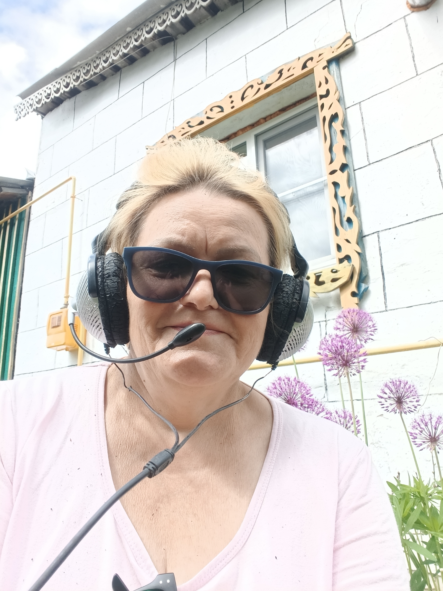 A medium shot captures a woman with blonde hair, wearing dark sunglasses and a headset with a microphone. She is positioned in front of a white brick building that has a decorative wooden window frame. The location appears to be outdoors, with blooming purple Allium flowers visible on the right side of the frame. A yellow gas pipe is seen on the building. The image was taken in Красный Маяк, Russia.