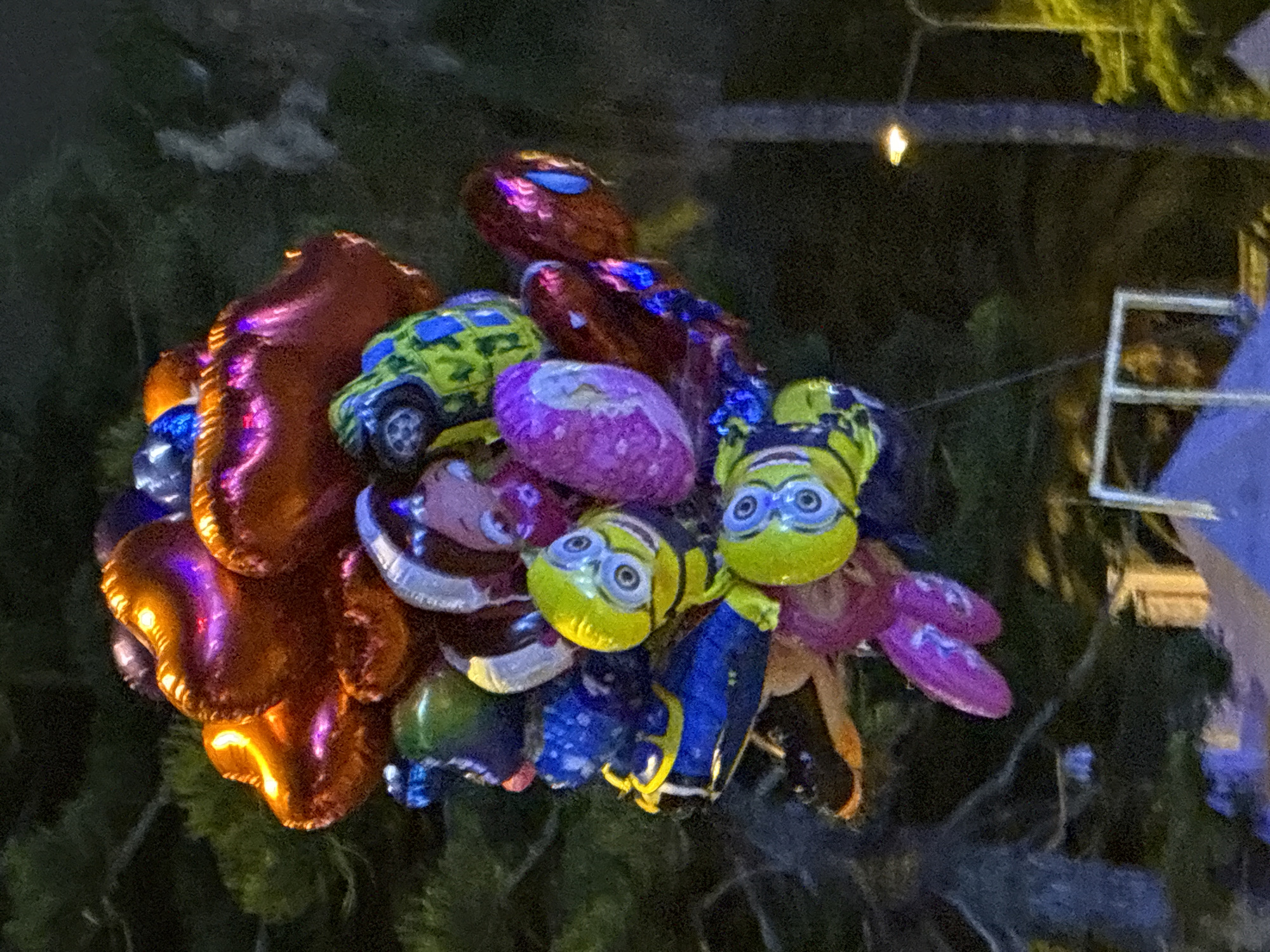 A cluster of colorful balloons is held together, some shaped like characters and objects. Prominently featured are several Minion-shaped balloons with their characteristic goggles and yellow bodies, some wearing blue overalls. There's also a balloon shaped like a green and yellow toy car. Other balloons are heart-shaped and metallic, appearing in shades of orange, purple, and blue. The balloons are illuminated by artificial light, which casts colorful reflections on their surfaces, suggesting an evening or nighttime setting. The background is dark and somewhat blurry, appearing to be foliage, possibly trees or bushes, with some hints of structures or lights in the upper right. The overall impression is of a festive arrangement of balloons, likely at an outdoor event or market. There is no visible text in the image.