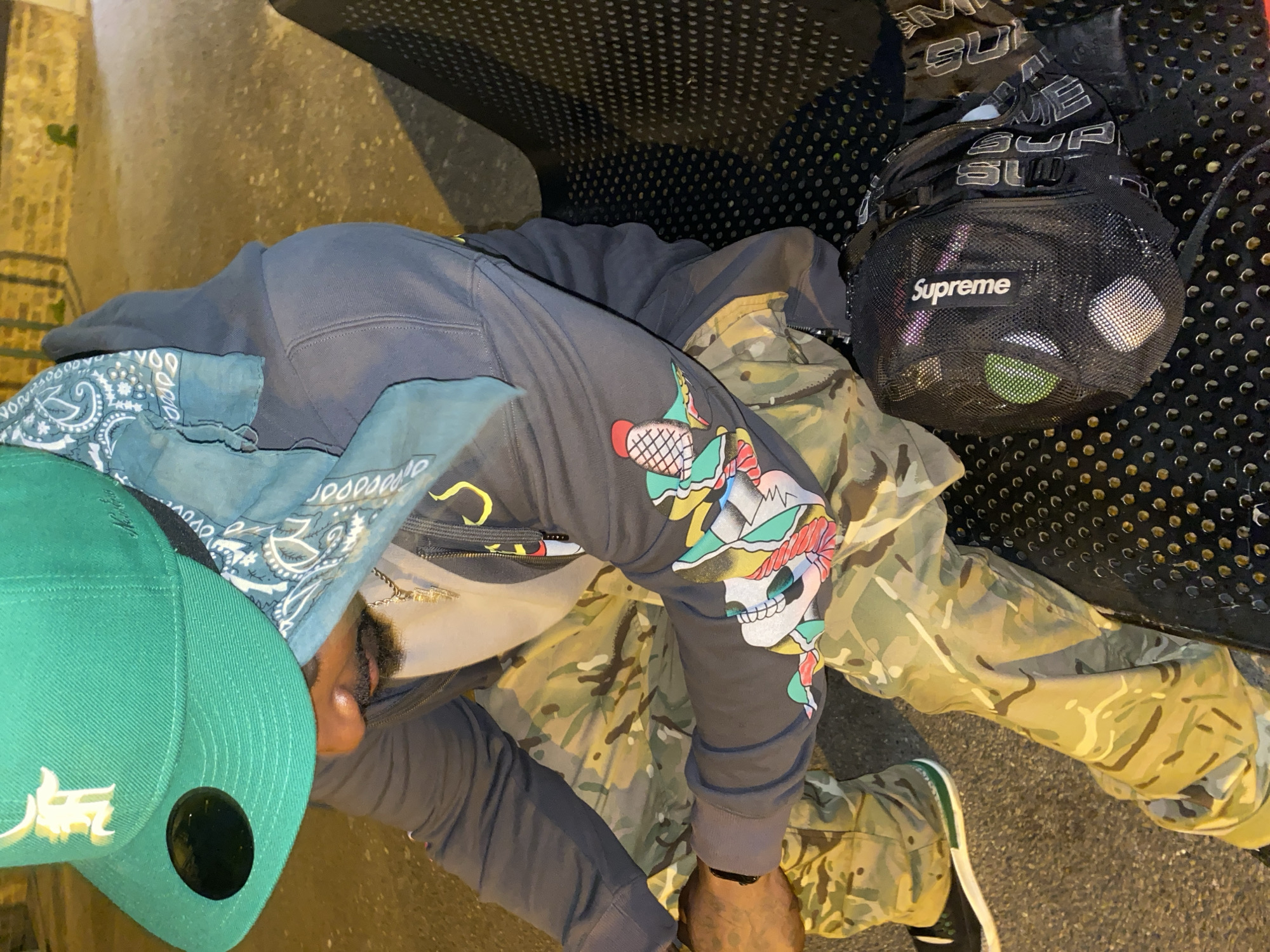 A man is sitting on a bench. He is wearing a green baseball cap, a bandana, a gray sweatshirt with a skull design on the sleeve, and camouflage pants. He is wearing black shoes. A black Supreme bag is sitting next to him on the bench. The bench is located in Waltham Cross, United Kingdom.