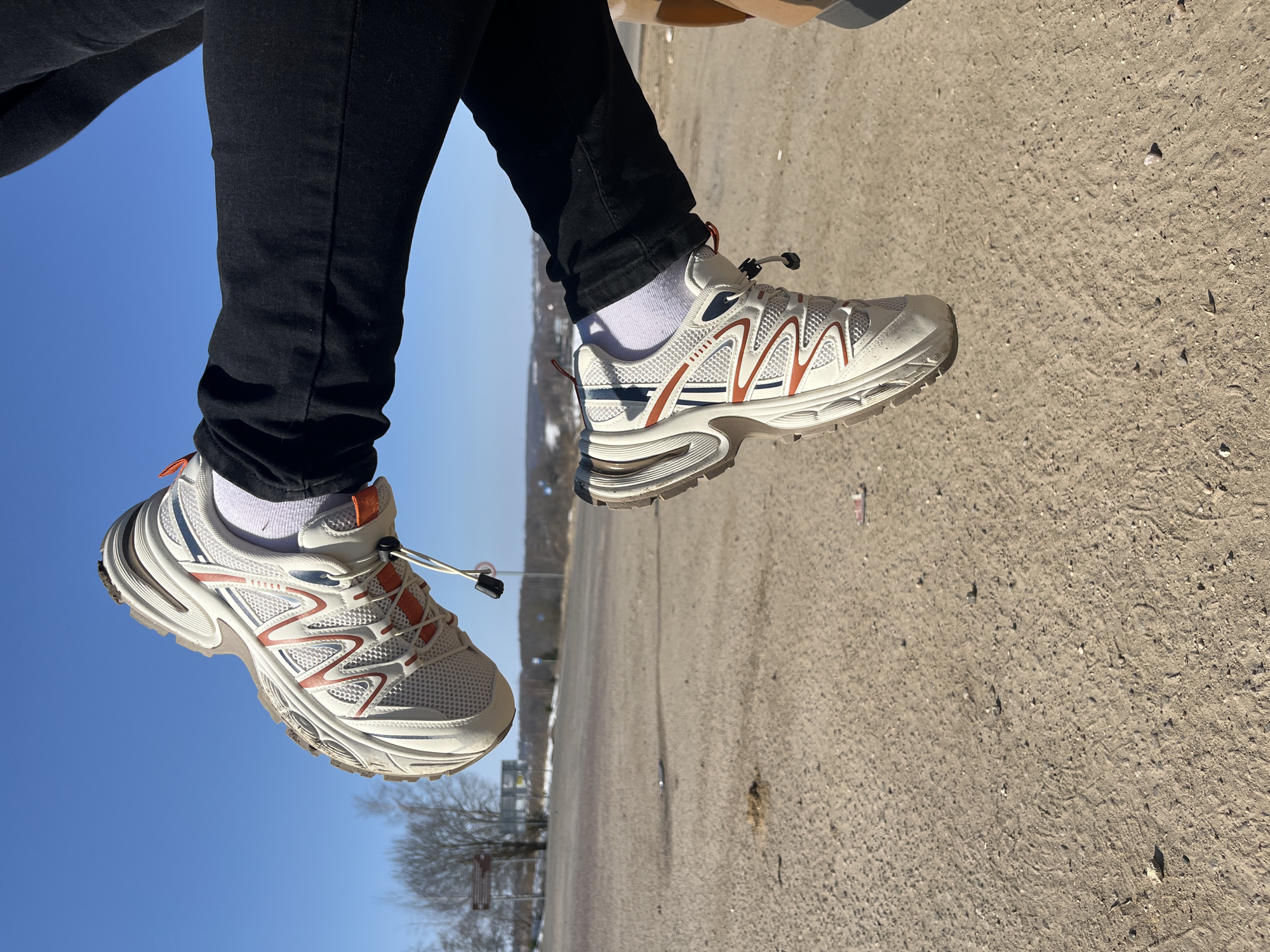 The image depicts the lower legs and feet of an individual, likely extending from a vehicle, set against an outdoor environment in Zubovo, Russia. The person wears black trousers, white ankle socks, and two light-colored athletic sneakers with white, beige, orange, and dark blue accents. The sneakers have textured soles and visible mesh panels. A portion of a dark-colored vehicle interior is visible on the right. Below the feet is a flat, unpaved ground composed of light brown soil and gravel, with scattered debris. In the mid-ground, bare deciduous trees and distant structures, including what appear to be road signs and utility poles, are visible. The background features a clear, bright blue sky.