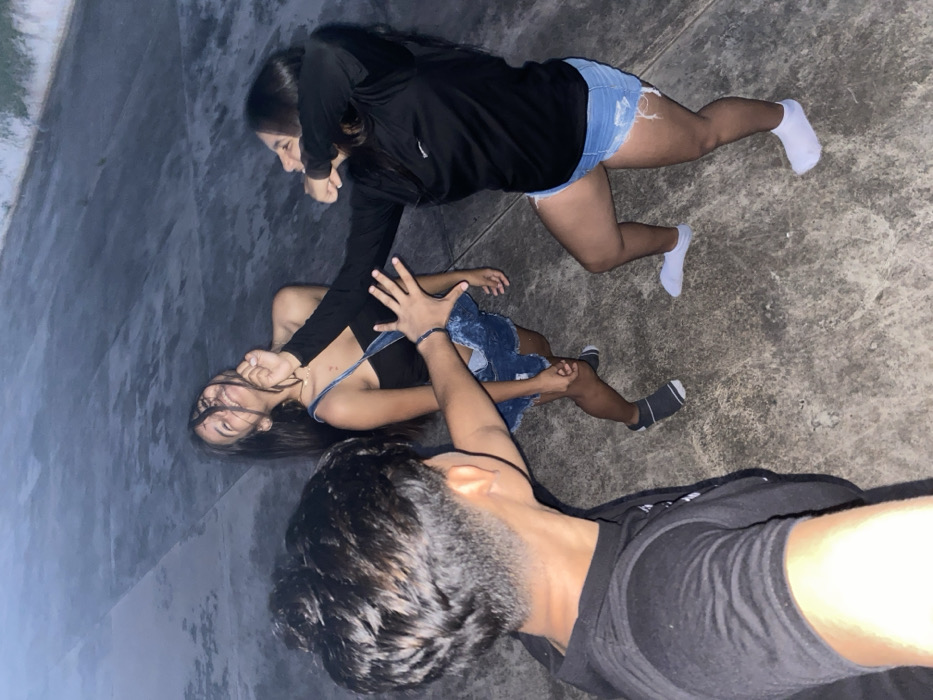 Three young individuals are depicted in an outdoor setting. One person in denim shorts and a black jacket stands over another person lying on the ground wearing denim overalls. The person standing appears to be play-fighting with the person on the ground. A third individual, wearing a black t-shirt is visible in the foreground, taking a selfie. The scene is dimly lit, and the ground appears to be concrete.
