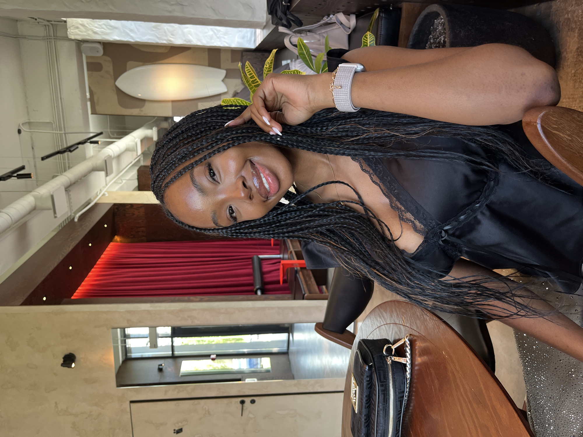 A medium shot captures a Black woman with braided hair sitting at a round wooden table in what appears to be a cafe or restaurant. She is looking at the camera and smiling, with her left hand resting on her cheek. She is wearing a black satin camisole with lace trim and a sparkly silver skirt. On the table in front of her is a black clutch purse. In the background, there is a red curtain, a white surfboard-like object mounted on the wall, and several potted plants. The wall texture is rough and beige, with some lights and electrical conduits visible on the ceiling. The general ambiance suggests an indoor, possibly trendy, establishment. The location of Kazan, Russia is not discernible from the visual information provided in the image.