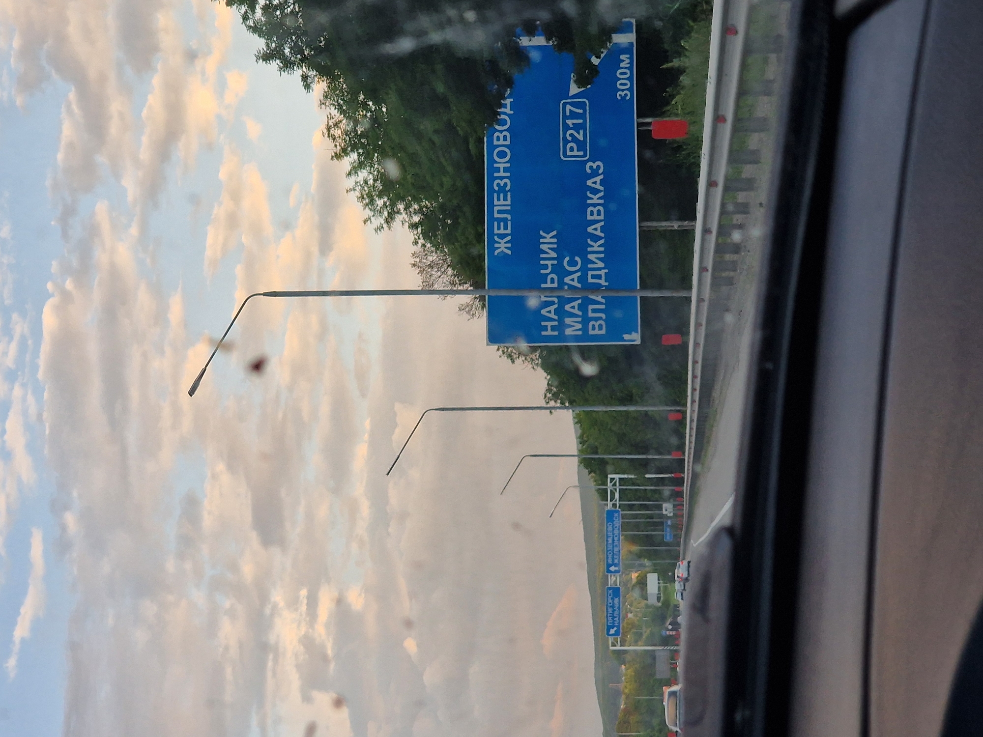 The image shows a highway scene with a large blue road sign in the foreground. The sign indicates destinations: "ЖЕЛЕЗНОВОДСК" (Zheleznovodsk) at the top, and below it "НАЛЬЧИК" (Nalchik), "МАГАС" (Magas), and "ВЛАДИКАВКАЗ" (Vladikavkaz). A route number "P217" and a distance "300M" are also visible on the sign. The road appears to be a multi-lane highway with a concrete barrier. Several streetlights are present along the roadside. In the background, there are trees and a cloudy sky. The perspective suggests the photo was taken from inside a vehicle. The presence of the town of Zmeyka is inferred by the road sign pointing to nearby cities such as Zheleznovodsk.