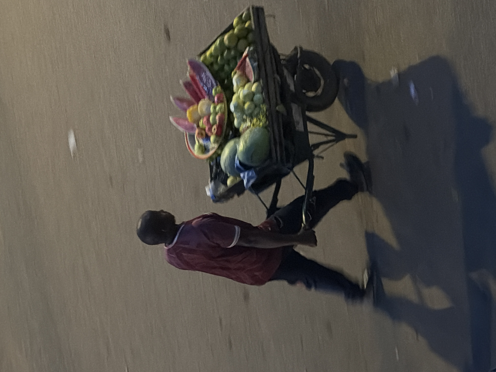A man pushes a fruit cart down a street in Bukuru, Nigeria. He is wearing a maroon t-shirt with white trim and dark pants. The cart is laden with a variety of fruits including watermelons, green apples, and what appear to be slices of watermelon and pineapple in a decorative arrangement. The cart has two wheels and a dark frame. The ground is a paved surface, possibly asphalt, with some small white debris scattered on it. Shadows are cast on the ground, indicating a light source from the right. The overall scene appears to be captured during daylight hours.