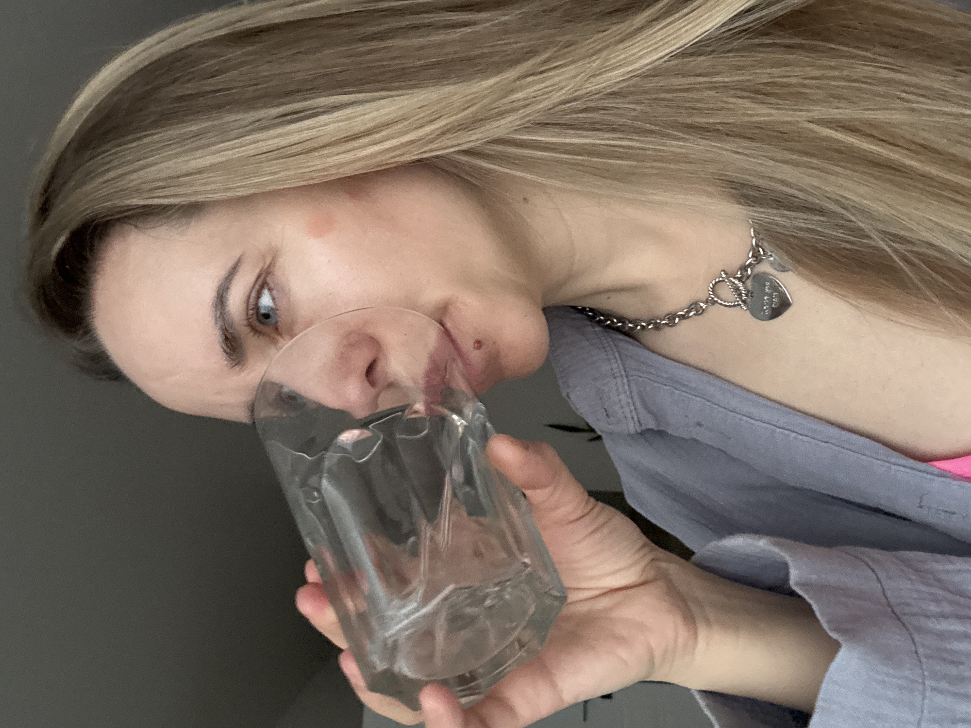 A woman with blonde hair is holding a clear glass with liquid inside. She is bringing the glass to her lips as if to drink. She has blue eyes, and a silver chain necklace with a heart-shaped pendant. She is wearing a blue shirt with a hint of a pink top visible underneath. The photo's background is blurry, making it difficult to determine the exact location, but there is no reliable data to confirm the location is in Chaikovsky, Russia.