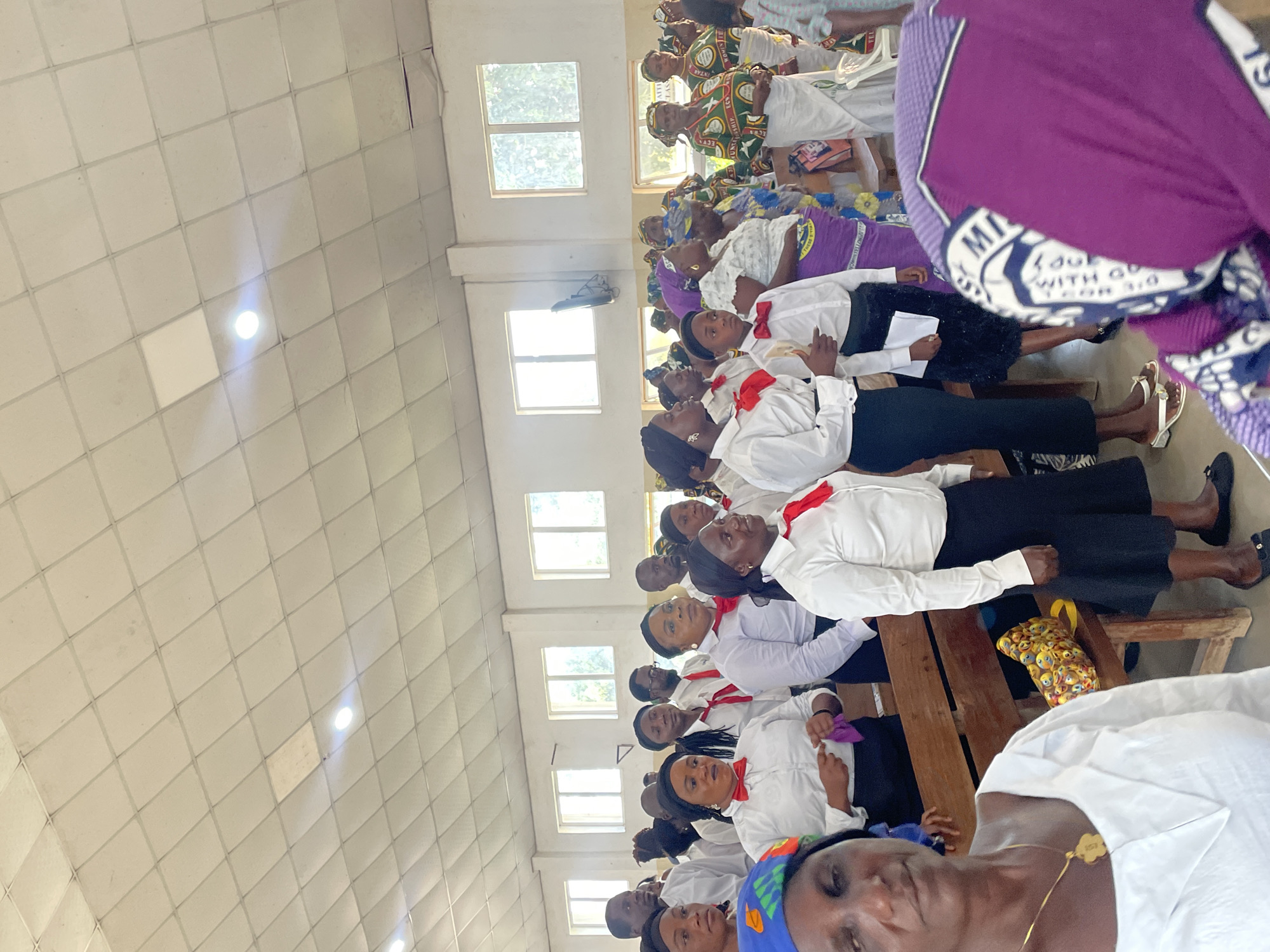 The photograph shows an interior scene filled with a gathering of people, likely in a church or assembly hall, located in Unknown, Unknown. A large group of individuals, primarily women, is seated on wooden benches. Many are wearing white shirts, black skirts, and red bow ties, suggesting a uniform or a specific group within the event. Some other women are wearing colorful headscarves and dresses. The room is brightly lit with artificial lights embedded in a tiled ceiling. There are windows visible, hinting at natural light sources as well. The overall atmosphere appears to be one of a formal gathering, potentially a religious service or a community event. The presence of printed materials in the hands of some people suggests possible reading or note-taking activities.