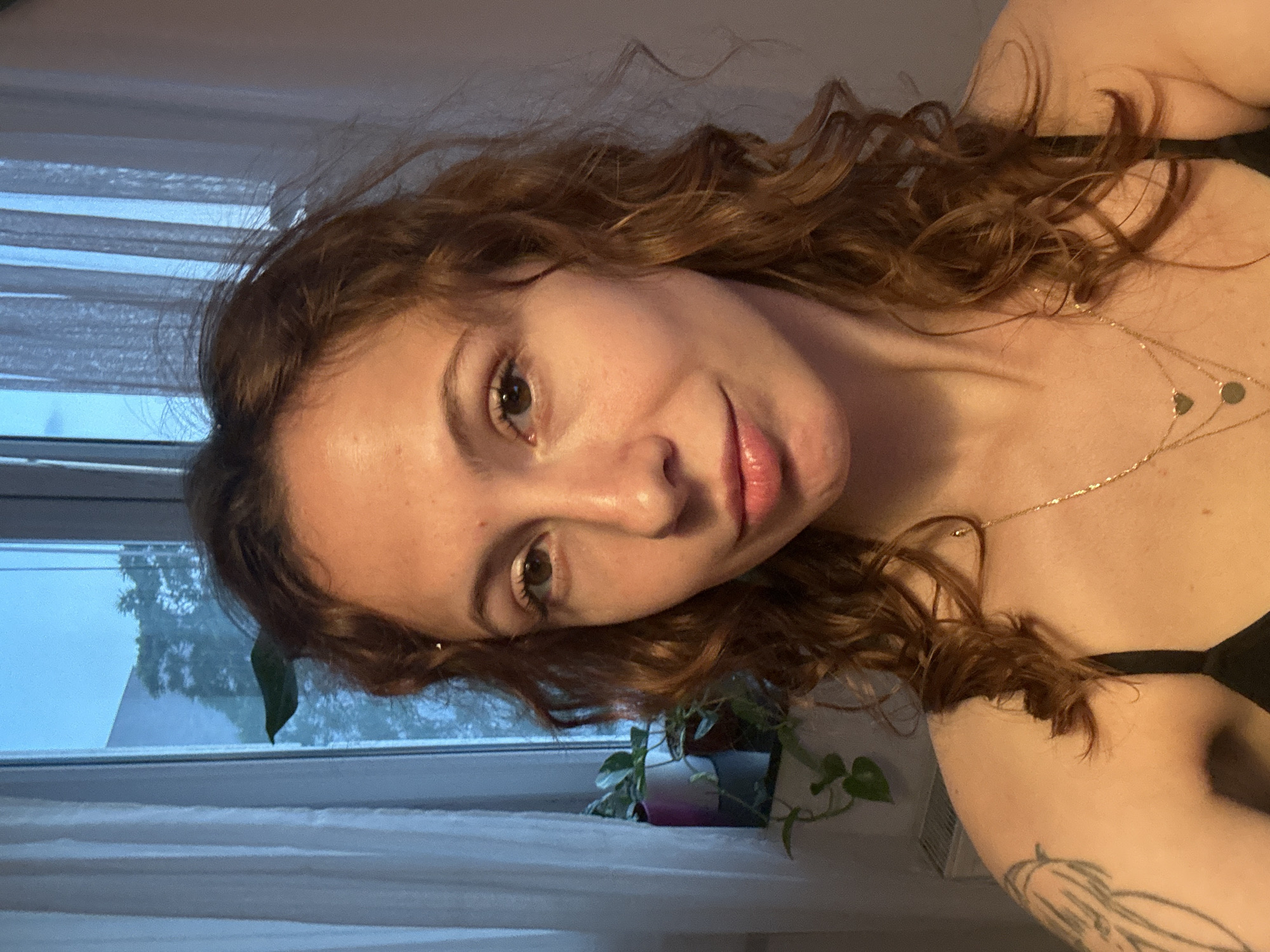A woman with curly reddish-brown hair is in the foreground, facing the camera with a slight smile. She is wearing a black top and a gold necklace with small pendants. A tattoo is visible on her left arm.
Behind her, a window with white blinds and a view of greenery outside is visible. A potted plant is on the windowsill.
There is no information in the image to identify the location as Novi Sad, Serbia.