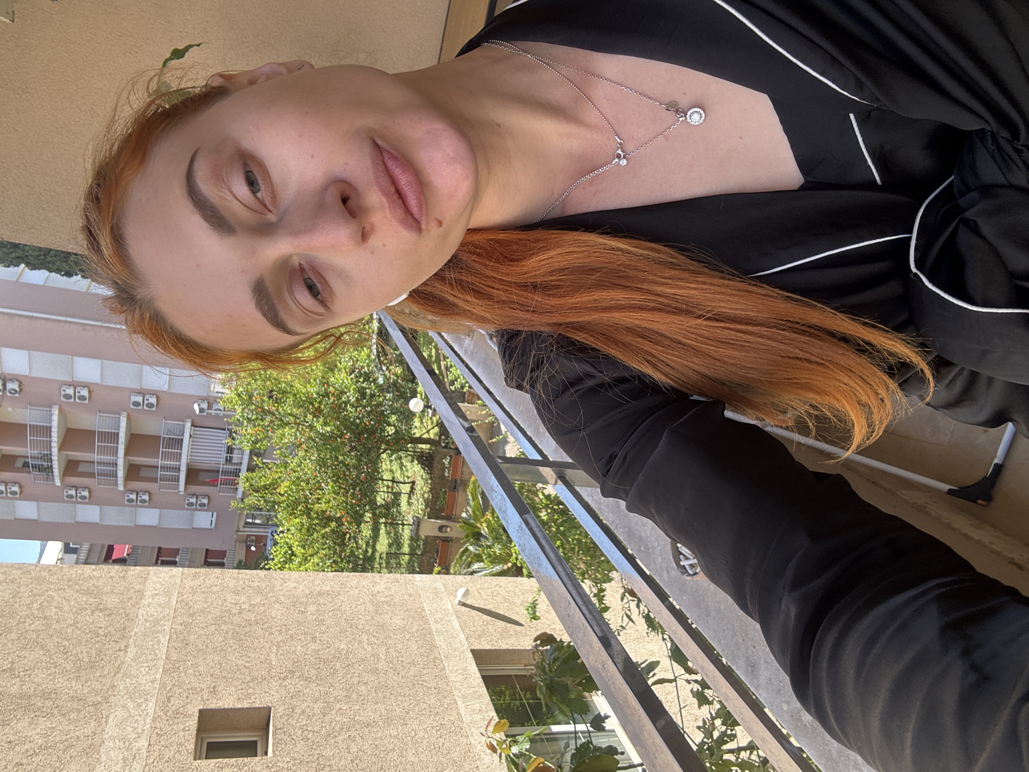 A medium shot captures a red-haired woman in a black pajama-like garment with white trim, positioned on a balcony. She is wearing a necklace with a circular pendant and a smaller pendant near the clasp. The balcony overlooks a garden with trees and flowers. In the background, a multi-story building with balconies and windows is visible. The location is likely within Budva, Montenegro, indicated by the architectural style and vegetation.