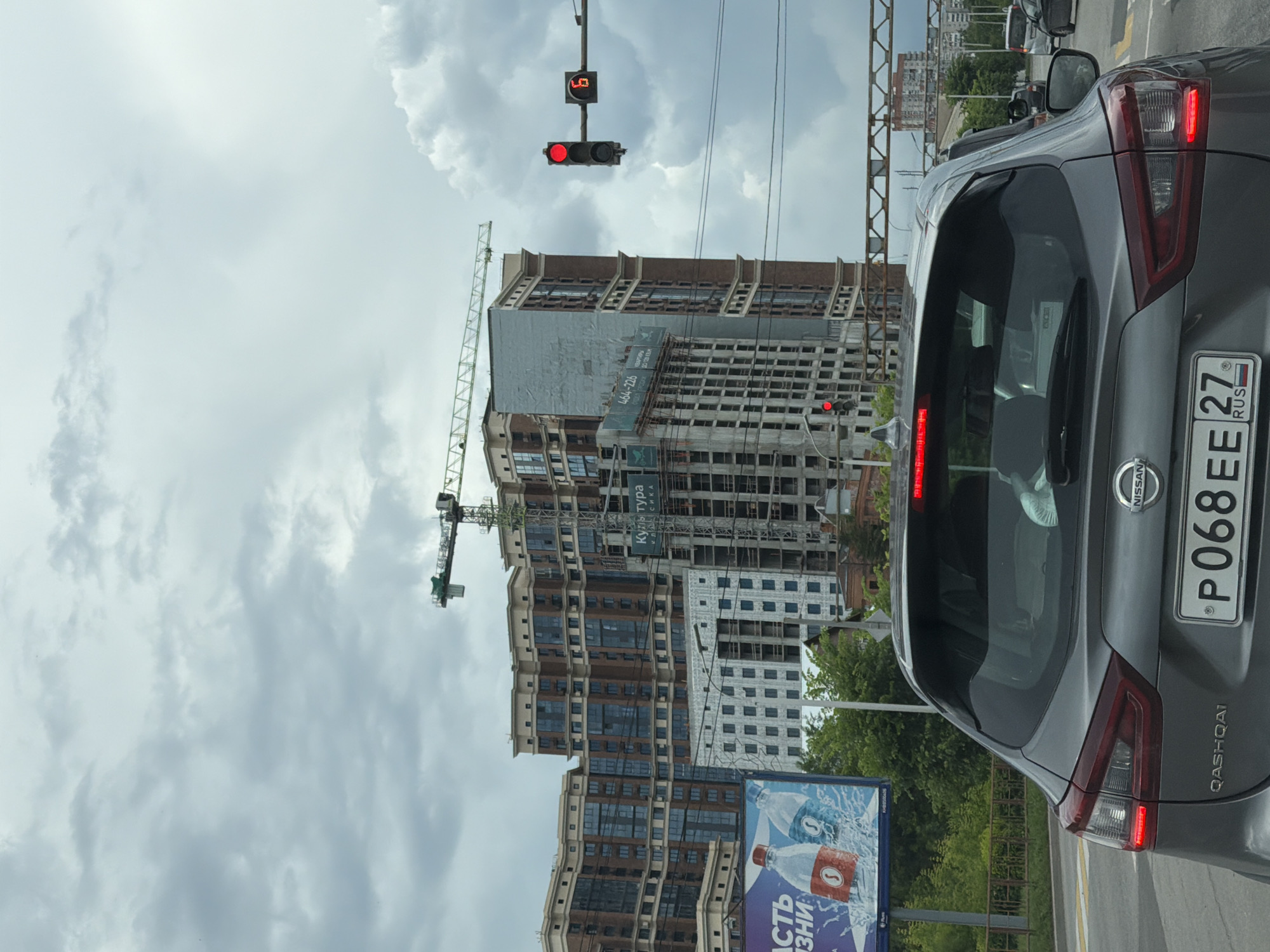 The image shows a grey Nissan Qashqai at a traffic light in Khabarovsk, Russia. The license plate reads "P068EE 27 RUS". In the background, there are several large, multi-story buildings under construction, with cranes visible. A billboard advertising bottled water is also present. The sky is overcast with clouds. The traffic light is red, indicating that the car is stopped. The image appears to be taken from another vehicle.