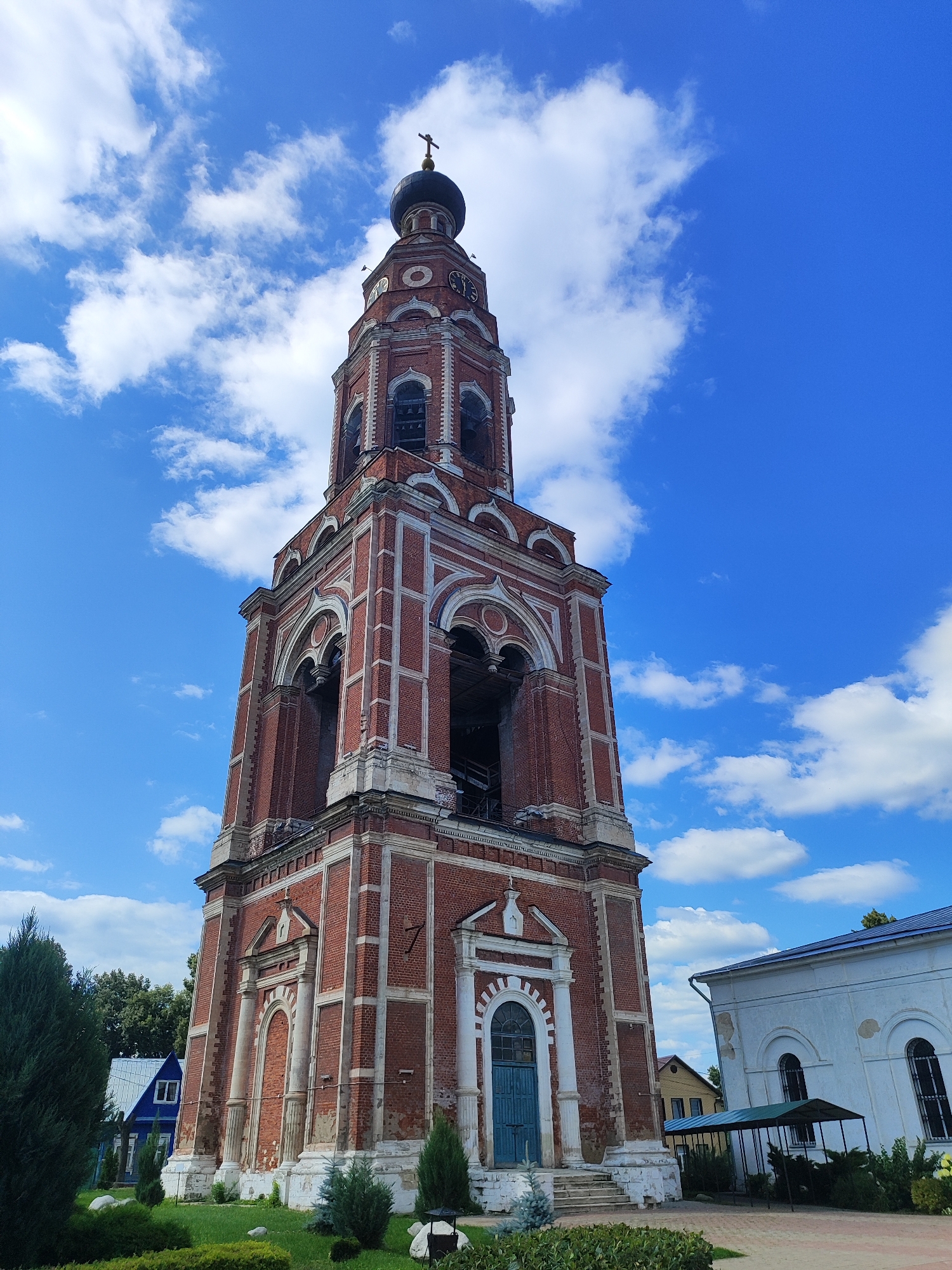 The image depicts a tall, red brick bell tower with white decorative elements, topped by a dark dome with a cross, under a bright blue sky with scattered white clouds. The bell tower features arched openings, some of which contain visible bells, and a clock face. To the right of the bell tower, a white building with arched windows and a blue roof is partially visible. In the foreground, there is a well-maintained green lawn with manicured bushes and small trees. To the left of the bell tower, a blue house with a white roof and a dark window is visible amidst lush green trees. The scene appears to be an exterior shot of a religious complex or historical site in Bronnitsy, Russia, likely a church or monastery, given the architectural style and presence of a bell tower. No people are present in the image.