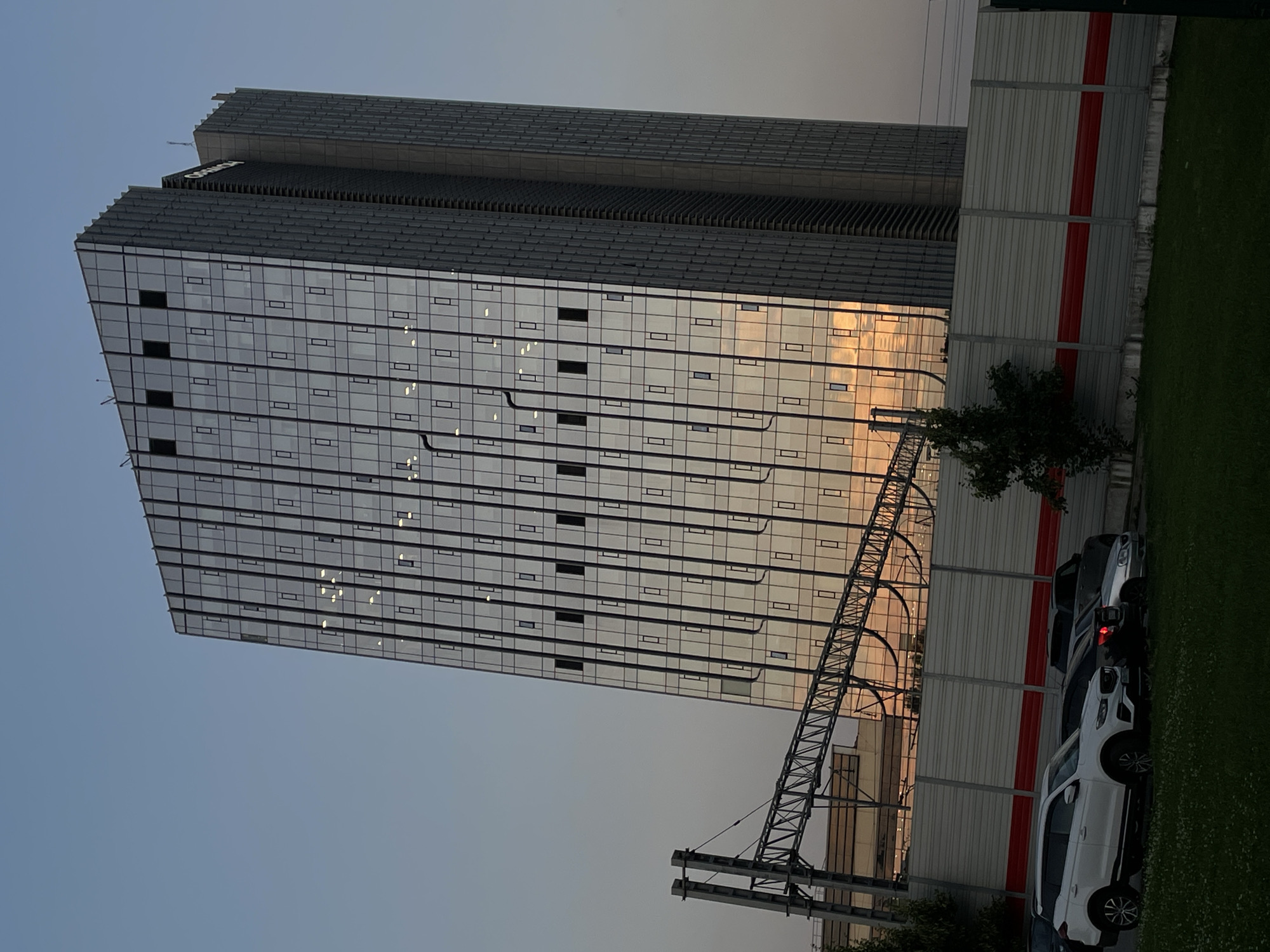 The image shows a modern skyscraper with a glass facade, reflecting the warm hues of a sunset or sunrise. The building has a series of horizontal window panes, with some lights visible inside, suggesting occupancy. The top of the skyscraper features a darker, tiered structure, and there's visible text on this upper section.
In the foreground, a metal framework structure, possibly part of an elevated railway or infrastructure, extends across the scene. To the right of the skyscraper, there's a light-colored barrier with two red horizontal stripes, bordering a well-maintained green lawn. Several cars are parked along the barrier, including a white SUV and other dark-colored vehicles.
The sky is a pale blue, transitioning into warmer tones near the horizon, indicative of either dawn or dusk. The overall atmosphere is serene, with the architecture of the building being the dominant subject. The presence of the cars and the manicured lawn suggest an urban or suburban setting, and the hints provided place the location in Trekhgorka, Russia. No specific activity or event is depicted, but the scene implies a peaceful end to the day or the beginning of a new one.