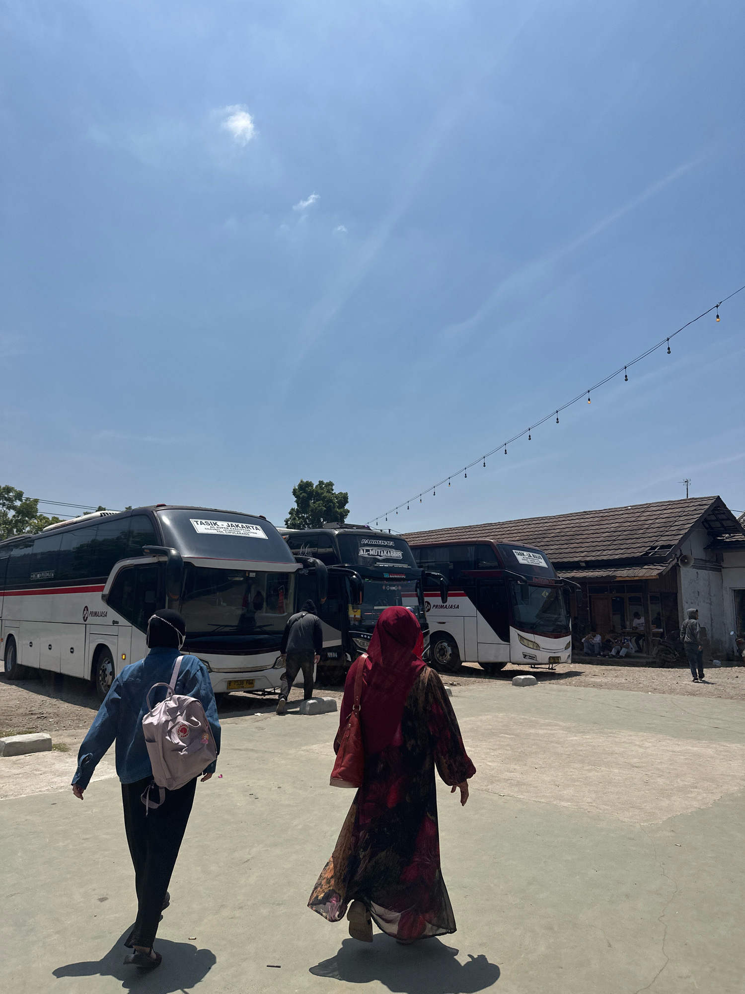 This image captures a daytime scene at a bus terminal or parking area in Garut, Indonesia. The foreground features two individuals walking away from the camera. The person on the left is wearing a blue denim jacket, black pants, and a light pink backpack. The person on the right is dressed in a dark red hijab and a long, patterned dress.
In the mid-ground, several large passenger buses are parked. One bus, predominantly white with red and black accents, displays the text "PRIMAJASA" and "TASIK-JAKARTA". Another bus, mostly black, also has visible signage. A weathered building with a tiled roof is situated in the background, with a group of people sitting and standing near its entrance. The ground is a mixture of paved surfaces and dirt. Overhead, a clear blue sky is adorned with a few wispy clouds and a string of decorative lights. The overall atmosphere suggests a travel hub on a sunny day.