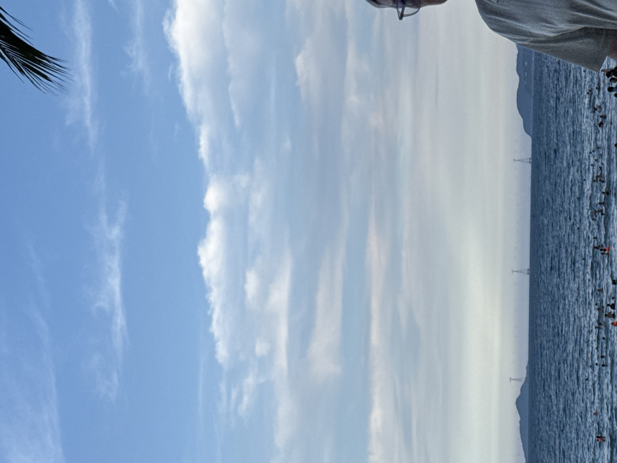 The image depicts a serene beach scene in Nha Trang Ward, Vietnam. In the foreground, a man wearing glasses and a light-colored shirt is partially visible, facing towards the right of the frame. His presence suggests he is an observer of the scenery. The middle ground is occupied by a vast expanse of calm, blue ocean, dotted with numerous people swimming and enjoying the water. Beyond the ocean, a mountainous island with a cable car system is visible in the distance. The sky above is a canvas of blue, adorned with scattered white clouds. The overall atmosphere is one of relaxation and enjoyment on a tropical beach.