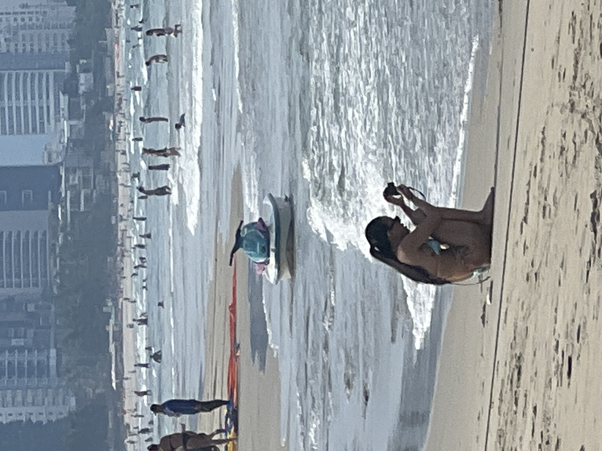 This image shows a beach scene in Đà Nẵng, Vietnam. In the foreground, a person is sitting on the sand with their legs extended, holding a camera and appearing to take a photo. They are wearing a swimsuit. Behind them, the ocean waves are breaking on the shore. Further back, several people are visible on the beach and in the water, some with surfboards. In the background, tall buildings line the coastline, suggesting an urban beach setting. The water appears bright and there are reflections on the surface, indicating a sunny day. The sand is light brown and there are some shadows, but the overall lighting suggests it is daytime, likely late morning or afternoon. The image is taken from a slightly elevated angle, looking down towards the beach and the water. There is no visible text in the image. The scene conveys a relaxed atmosphere of a popular beach destination.