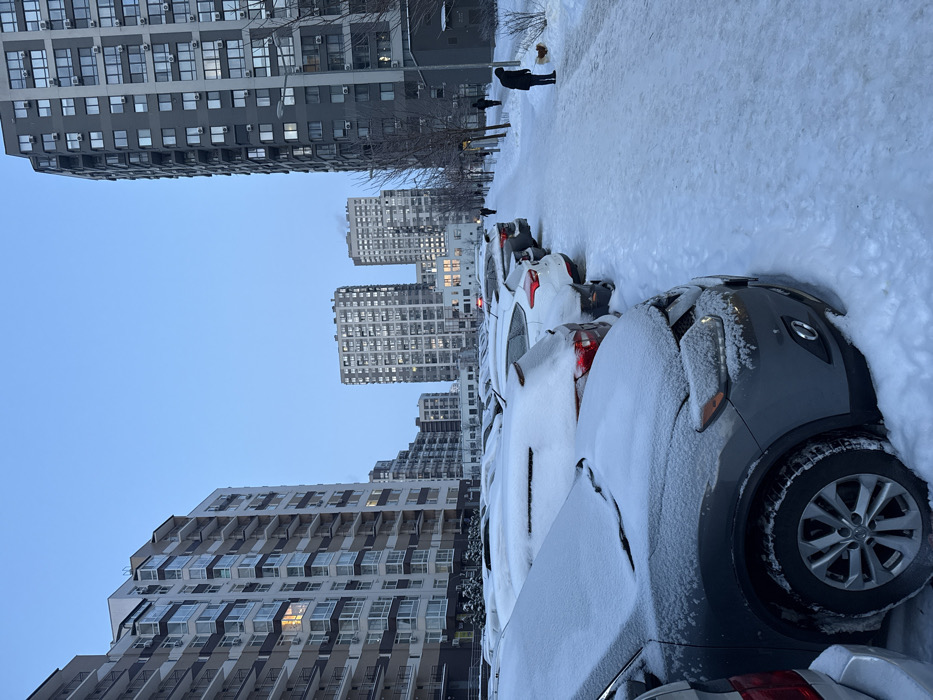 A winter scene with parked cars covered in snow. High-rise buildings are in the background under a blue sky. People walk on a snowy path alongside the cars. The focus is on the snow-covered vehicles and urban architecture.