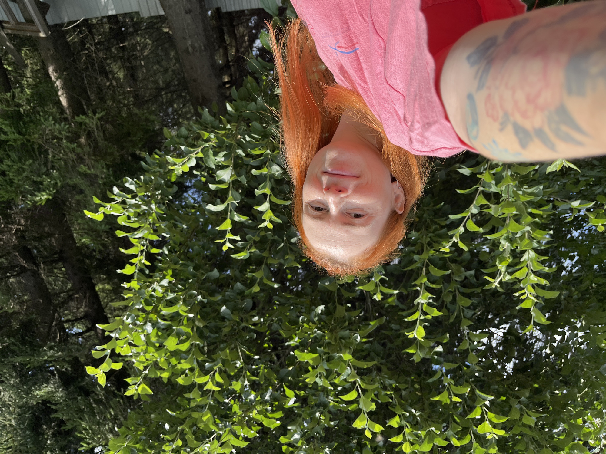 A low-angle, medium shot captures a woman with bright red hair holding a camera up to take a selfie. She is wearing a pink t-shirt and has tattoos on her left arm and shoulder. Behind her, a dense canopy of green leaves creates a vibrant backdrop, with the dappled sunlight filtering through. The woman is smiling and looking towards the camera. The scene appears to be outdoors, possibly in a garden or park, with the lush foliage suggesting a natural setting. The specific location in Igra, Russia, cannot be determined from the visual information provided in the image.