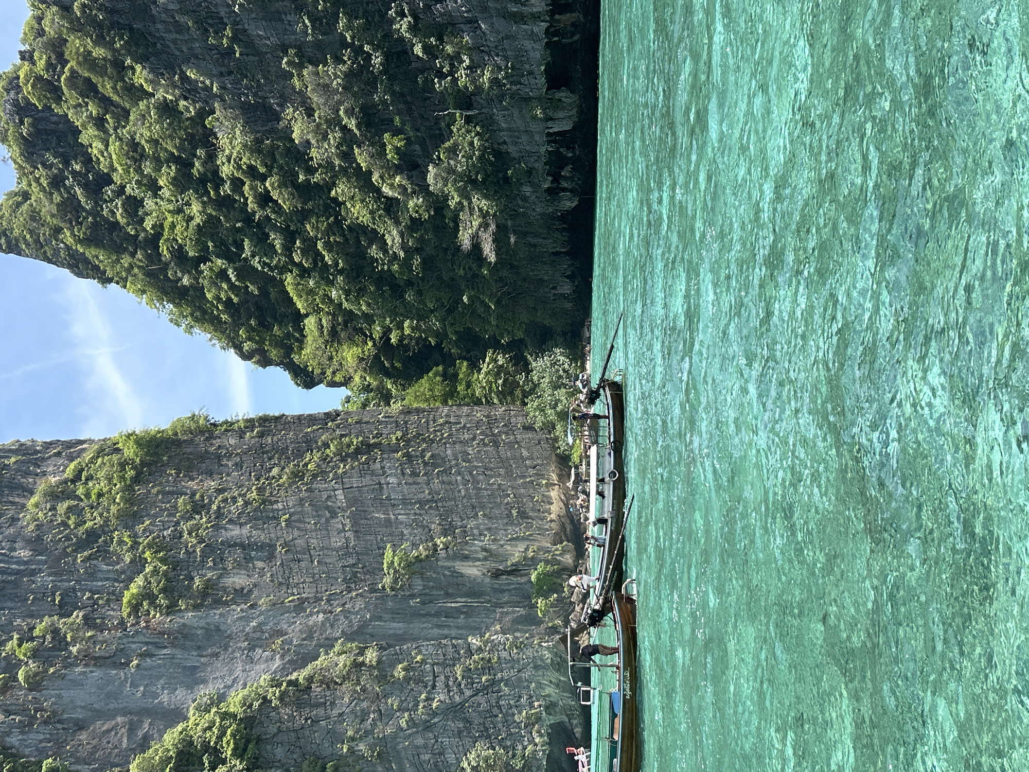 The image shows a view of the turquoise waters of Ao Nang, Thailand, with two longtail boats in the foreground. The boats are filled with people. Towering limestone cliffs covered in lush green vegetation rise dramatically from the water on either side of the scene. The sky is clear and blue with a few wispy clouds. The sun is shining, casting light on the water and cliffs. The water is clear and reveals the sandy bottom beneath. One boat has a canopy for shade and appears to be moving. The other boat is anchored or moving slowly. The overall impression is a beautiful tropical paradise.