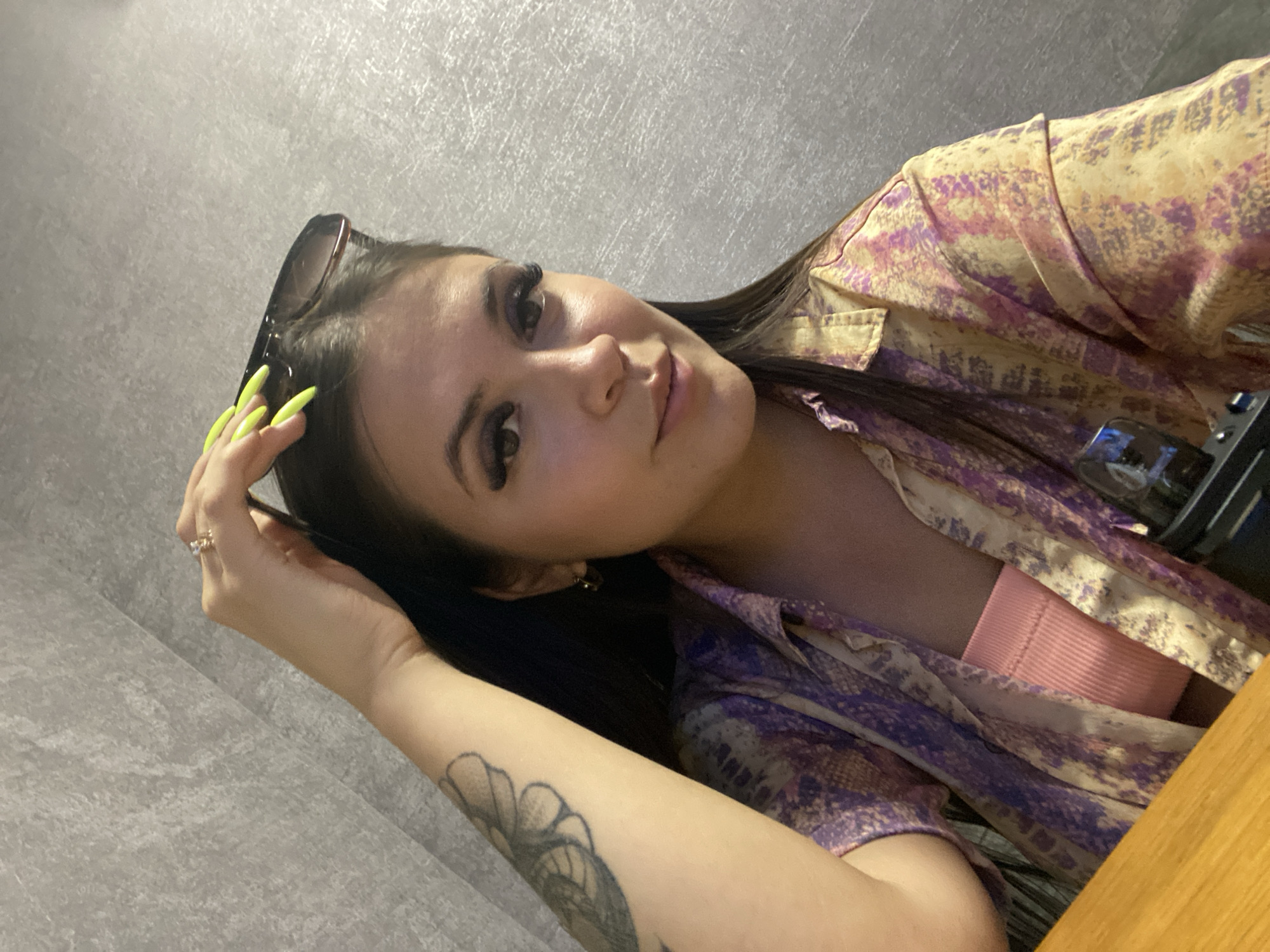 The image shows a young woman with dark hair, long eyelashes, and bright yellow nails. She is wearing a pink top and a colorful, patterned button-down shirt. She is touching her hair, which has sunglasses resting on top of it. A tattoo is visible on her left arm. She is wearing a ring on her right hand. The background is a gray, textured wall. The image appears to be taken indoors, possibly in a restaurant or cafe, with a wooden surface visible in the foreground. There is no specific visual information to indicate the location is Makhachkala, Russia.