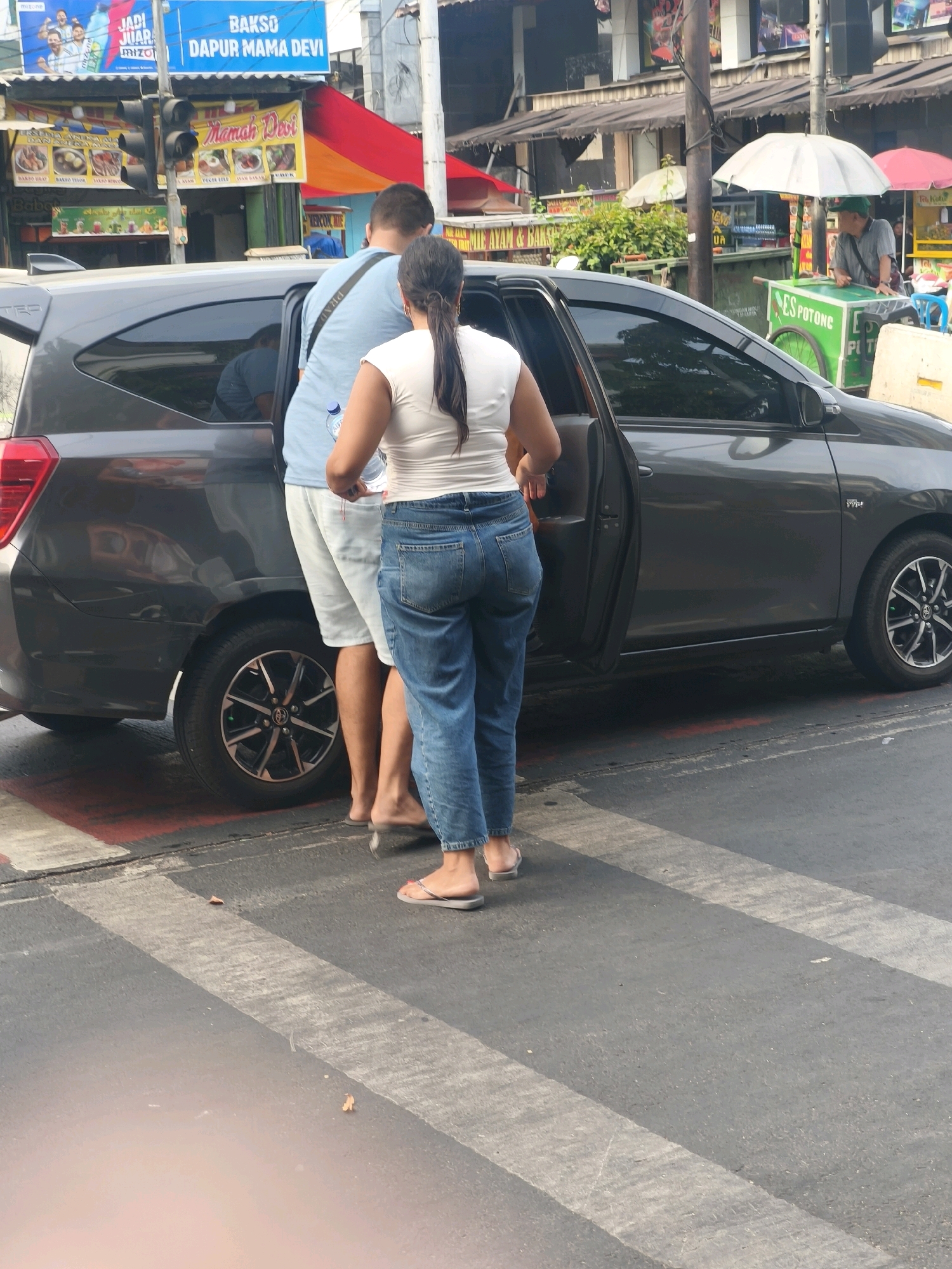 The image depicts a street scene in West Jakarta, Indonesia, likely near a food stall area. In the foreground, a dark gray car is parked with its driver's side door open. A man wearing a light blue t-shirt and white shorts stands next to the car, while a woman in a white top and blue jeans stands behind him, facing away from the camera. Both individuals are on a pedestrian crossing marked with white stripes. Behind the car, there are several food stalls. One stall features a red and yellow awning, with a banner that reads "BAKSO DAPUR MAMA DEVI" and displays images of food. Further back, another stall with a green cart and an umbrella is visible, with a person in a gray shirt and green cap tending to it. The environment suggests a bustling urban area with a focus on street food vendors.