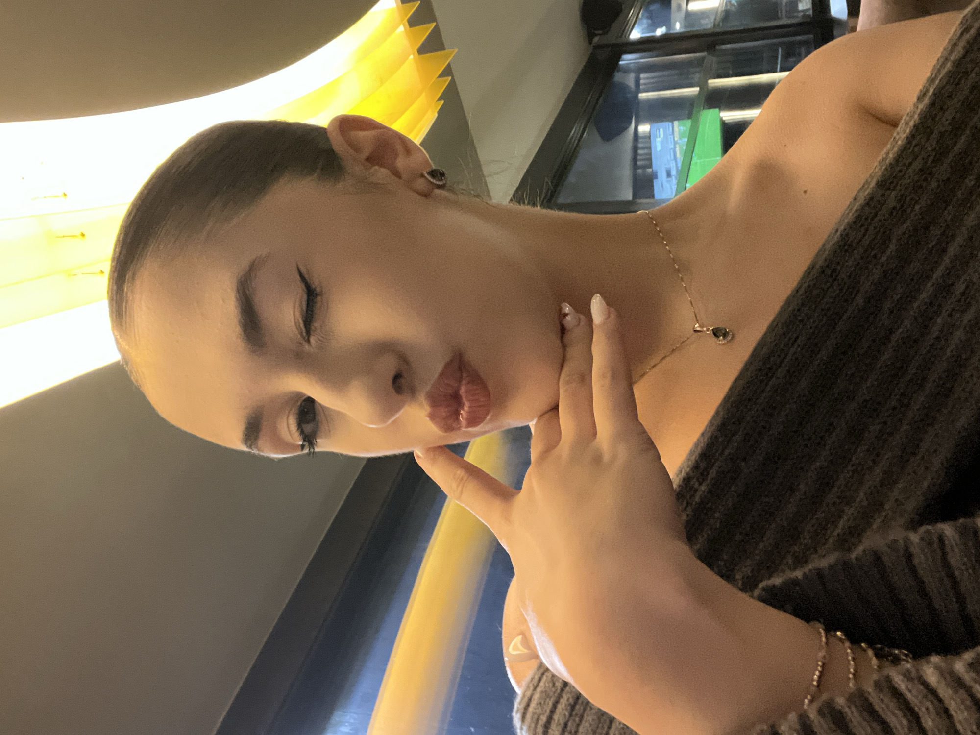 A young woman poses indoors. The lighting is mixed, with a combination of natural and artificial light creating both highlights and shadows on her face. The location cannot be determined based on the image.
**Subject:**
* A young woman with her hair pulled back and minimal makeup.
* She is making a pouting or kissing face toward the camera.
* She is wearing a dark grey sweater with a low neckline, exposing her shoulder and upper chest. A delicate gold necklace with a dark pendant is visible around her neck.
**Objects:**
* She has a gold bracelet on her wrist.
* Her left hand is resting on her chin with the index finger extended upward. Her nails are short and painted with a light color.
**Environment:**
* The background is blurred. A yellow light fixture.
* There are windows visible with some dark elements, possibly the reflection of the surroundings.
