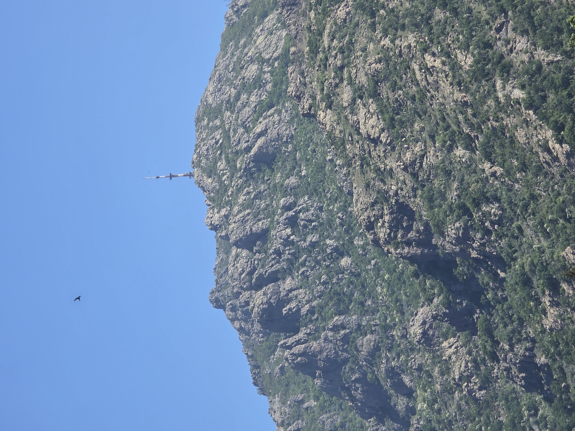 The image shows a rocky mountain face against a clear blue sky. A radio tower or antenna is visible on the edge of the mountain. A small bird is flying in the sky to the left of the mountain. The mountain is covered in rocky formations with some vegetation.