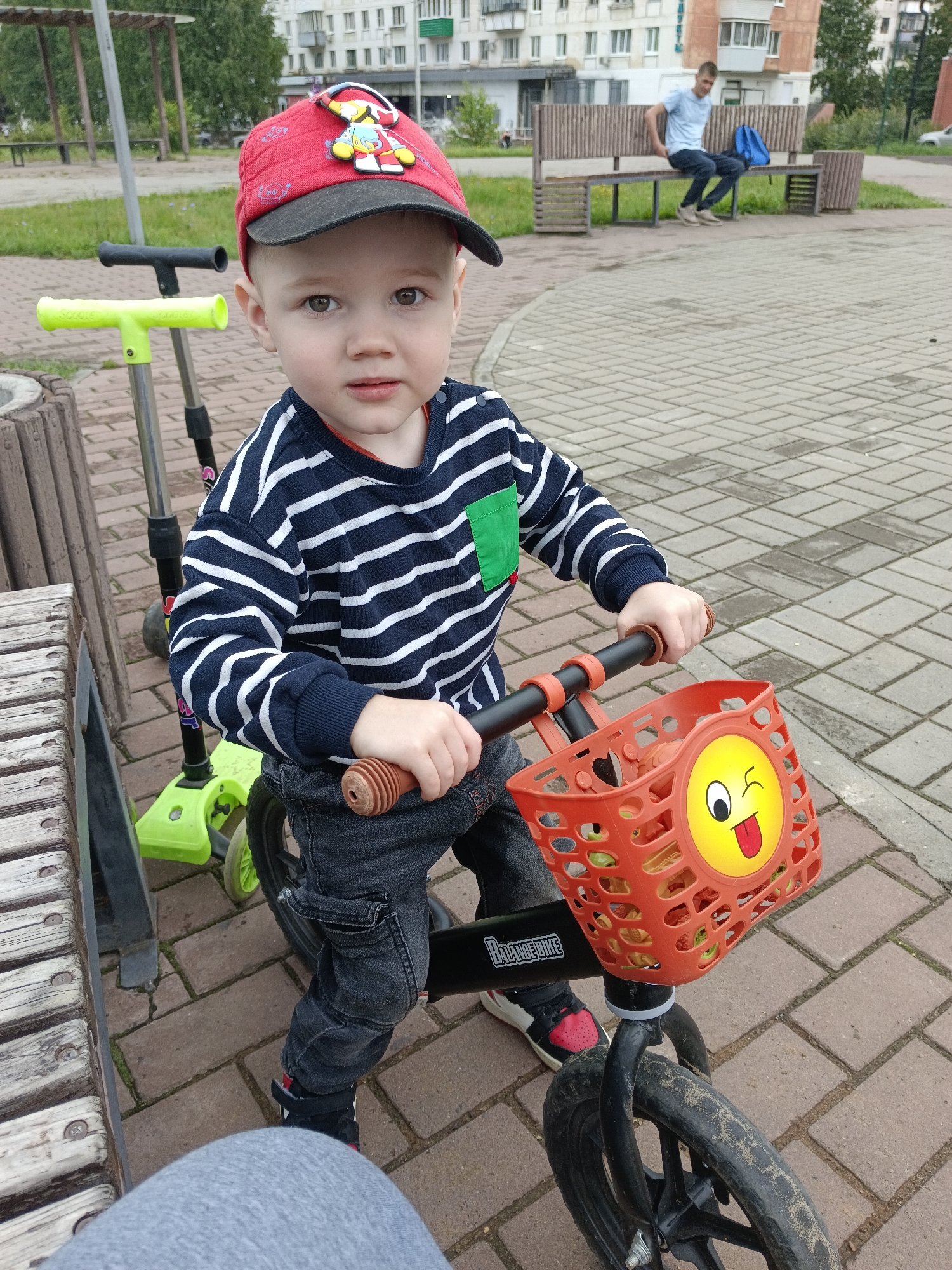 A young boy wearing a red baseball cap with a character on it and a navy and white striped long-sleeved shirt is sitting on a black balance bike. He is also wearing dark jeans and red and black sneakers. The balance bike has an orange basket on the front with a yellow smiley face sticker. Behind the boy, a green scooter is visible. The boy is on a paved pathway made of reddish-brown bricks. To the left, a wooden bench is partially visible. In the background, a man is sitting on a wooden bench, and a blue backpack is beside him. There are buildings and trees in the far background, consistent with a residential area in Chaikovsky, Russia. The overall setting appears to be an outdoor public space.