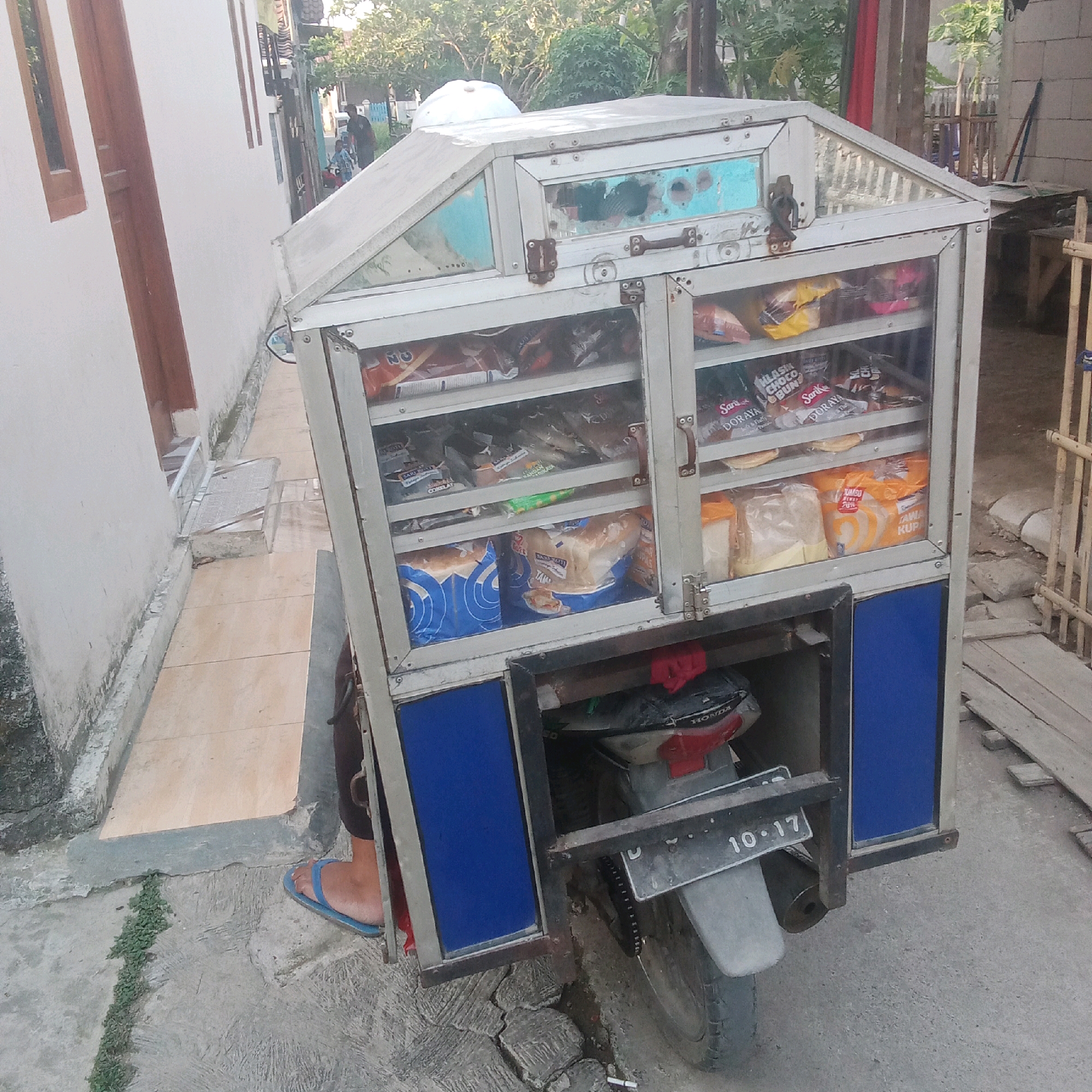 The image depicts a mobile food cart, likely a bread cart, attached to a motorcycle. The cart is constructed of metal and glass, with several shelves displaying various packaged food items, primarily bread loaves and snacks. The cart has a distinctive blue panel on its front lower section. A license plate that reads "10.17" is visible on the back of the motorcycle. A person's leg, wearing a blue flip-flop, is visible near the bottom left of the frame, suggesting the cart is being operated or has recently been parked.
The environment appears to be a residential or urban street in Karawang, Indonesia. The background includes a building with white walls and wooden window frames, trees, and other structures that suggest a typical Indonesian street scene. The ground is paved with concrete and tiles, with some patches of grass. The lighting indicates it is daytime, possibly late afternoon given the angle of the shadows. The overall scene is utilitarian, focused on the commercial aspect of the food cart.