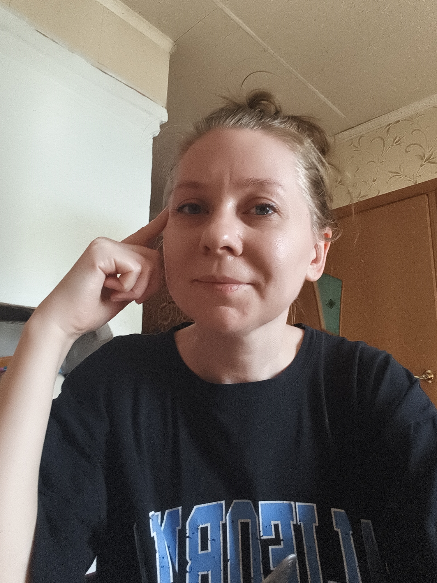 A woman is pictured indoors, resting her right temple with her right hand. She is wearing a black t-shirt with a blue and white logo. Her hair is pulled up in a bun. The background includes a wooden door, a wall with floral wallpaper, and a white ceiling with trim. The lighting suggests an indoor setting, possibly a room in a house. No specific location in the city of Ilmino, Russia can be determined from the image.