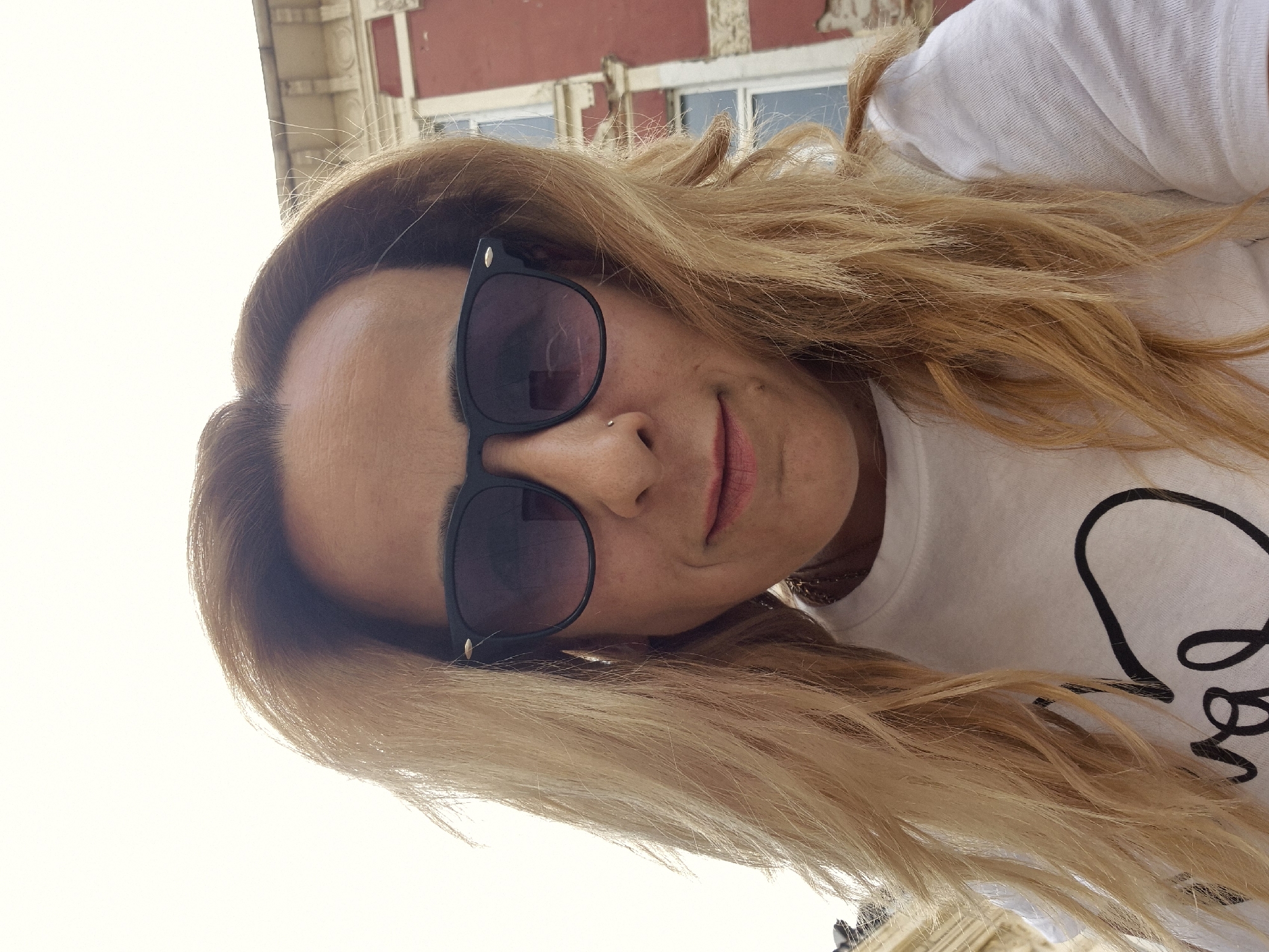 A selfie of a person with long, blonde hair, wearing sunglasses and a white t-shirt, with a building visible in the background. The location is the city of Unknown, Unknown.
