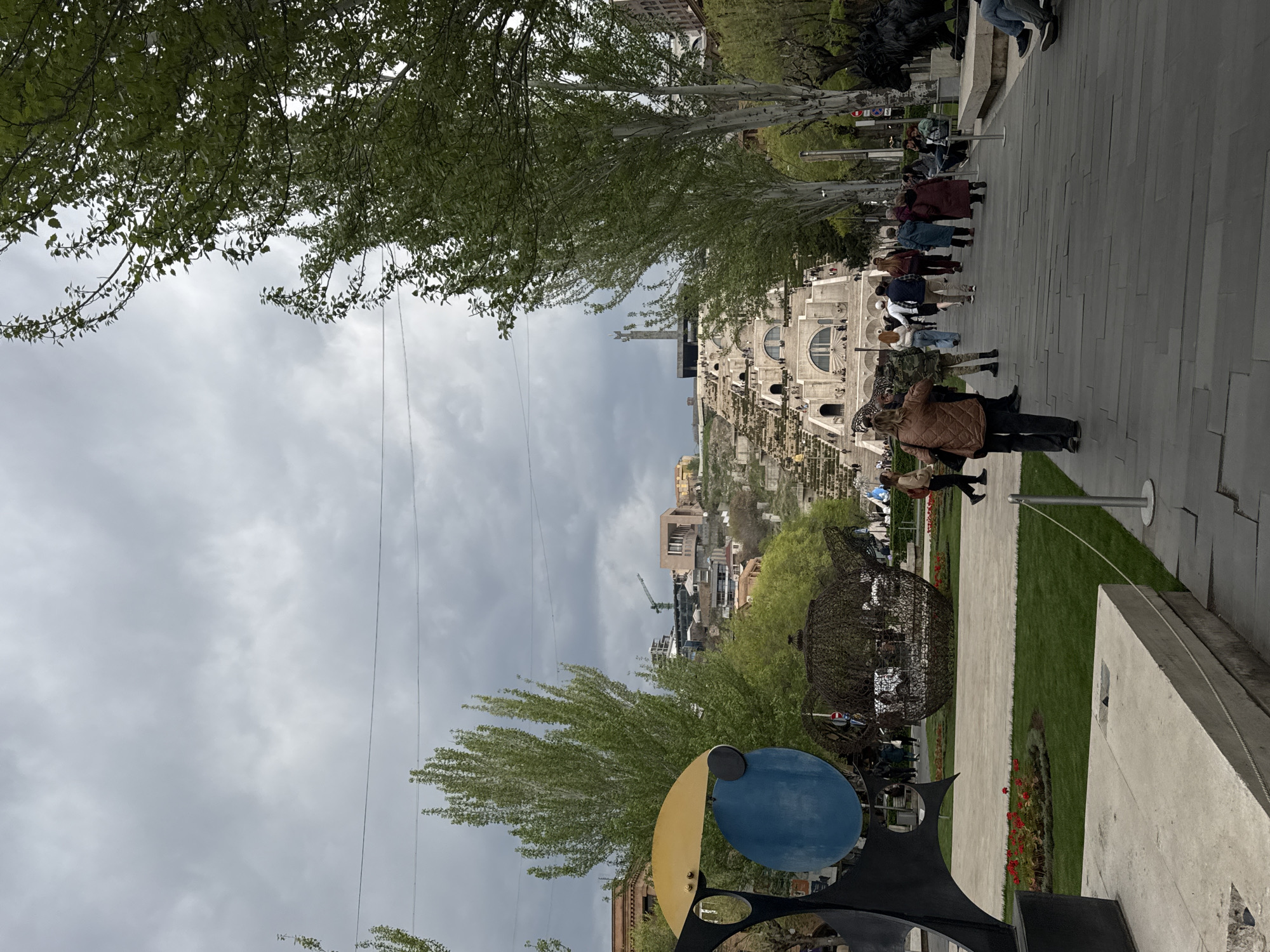 An outdoor scene captures a public park or plaza in Yerevan, Armenia, on an overcast day. The image is oriented vertically, but described as if correctly rotated.
In the mid-ground, the grand tiered architecture of the Yerevan Cascade dominates, featuring multiple levels of steps and semicircular windows or arches. At the summit of the Cascade, a tall monument, likely the Mother Armenia statue, is visible against the cloudy sky. Beyond the Cascade, hills dotted with urban buildings, including some under construction with a visible crane, form the background.
The foreground features a wide paved walkway on the right, made of grey rectangular tiles, where numerous people are strolling. Many individuals and small groups are seen walking up the path, dressed in light to medium jackets, suggesting mild weather. One person in a brown quilted jacket is prominent on the right, appearing to take a photograph.
On the left side of the walkway, lush green trees with abundant leaves indicate spring or early summer. Below the trees, a vibrant abstract sculpture with large blue and yellow circular elements stands prominently in the bottom left. Further along the grassy area to the right, another large, dark, intricately designed sculpture, possibly made of woven metal, is visible. Patches of green grass with small red flowers (likely tulips) add splashes of color to the landscape. Overhead, several power or communication lines stretch across the cloudy sky. The overall scene depicts a leisurely atmosphere in a significant cultural landmark. No legible text is visible.