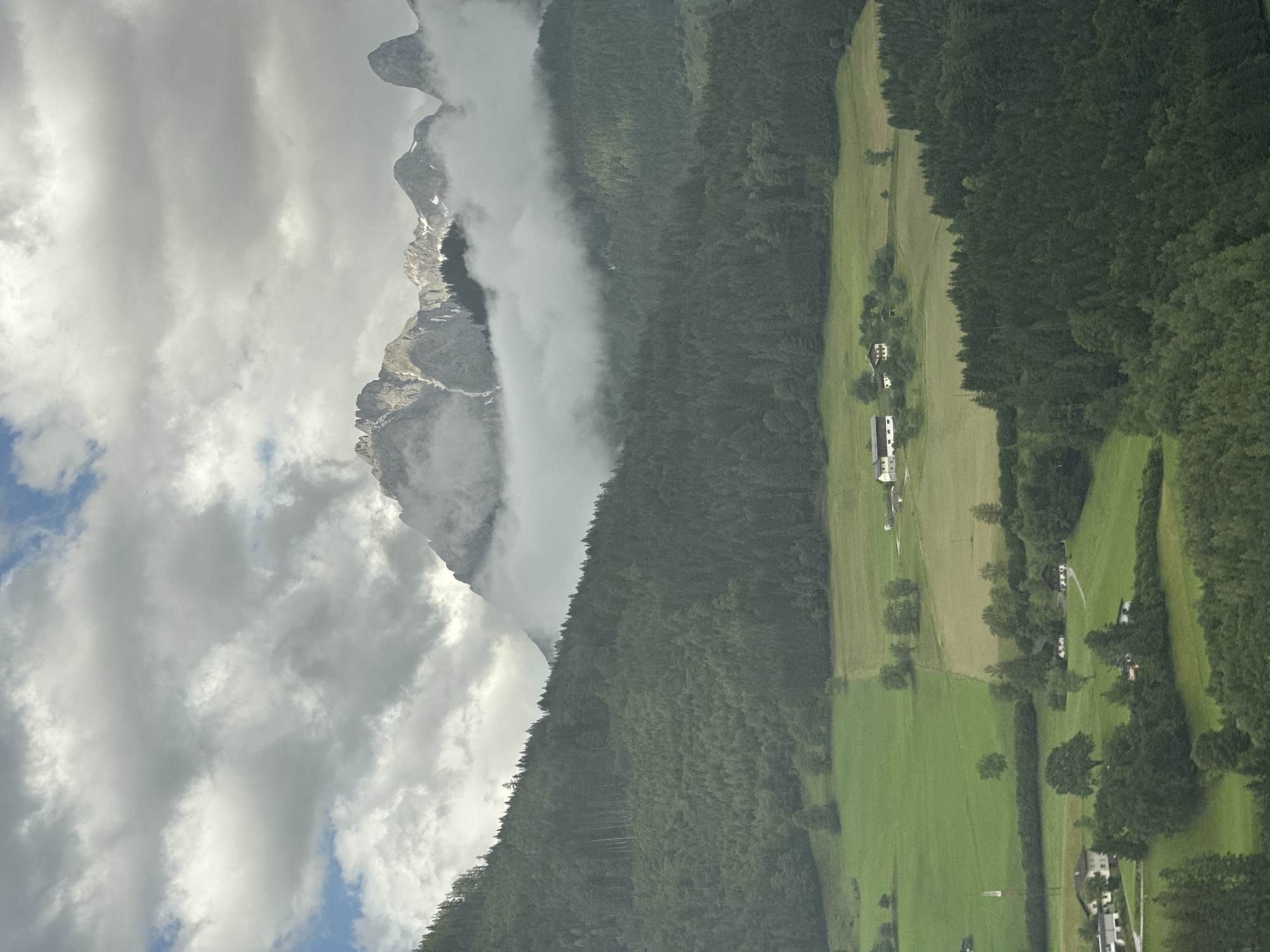 This image captures a majestic mountain landscape in Bischofshofen, Austria, under a dramatic cloudy sky. The foreground features rolling green hills dotted with scattered trees and a few buildings, suggesting rural settlements. A dense forest covers the slopes of the hills, transitioning into the towering mountain range in the background. The peaks of the mountains are partially obscured by a thick layer of clouds and mist, with snow patches visible on some of the higher elevations. The overall scene conveys a sense of natural grandeur and tranquility.