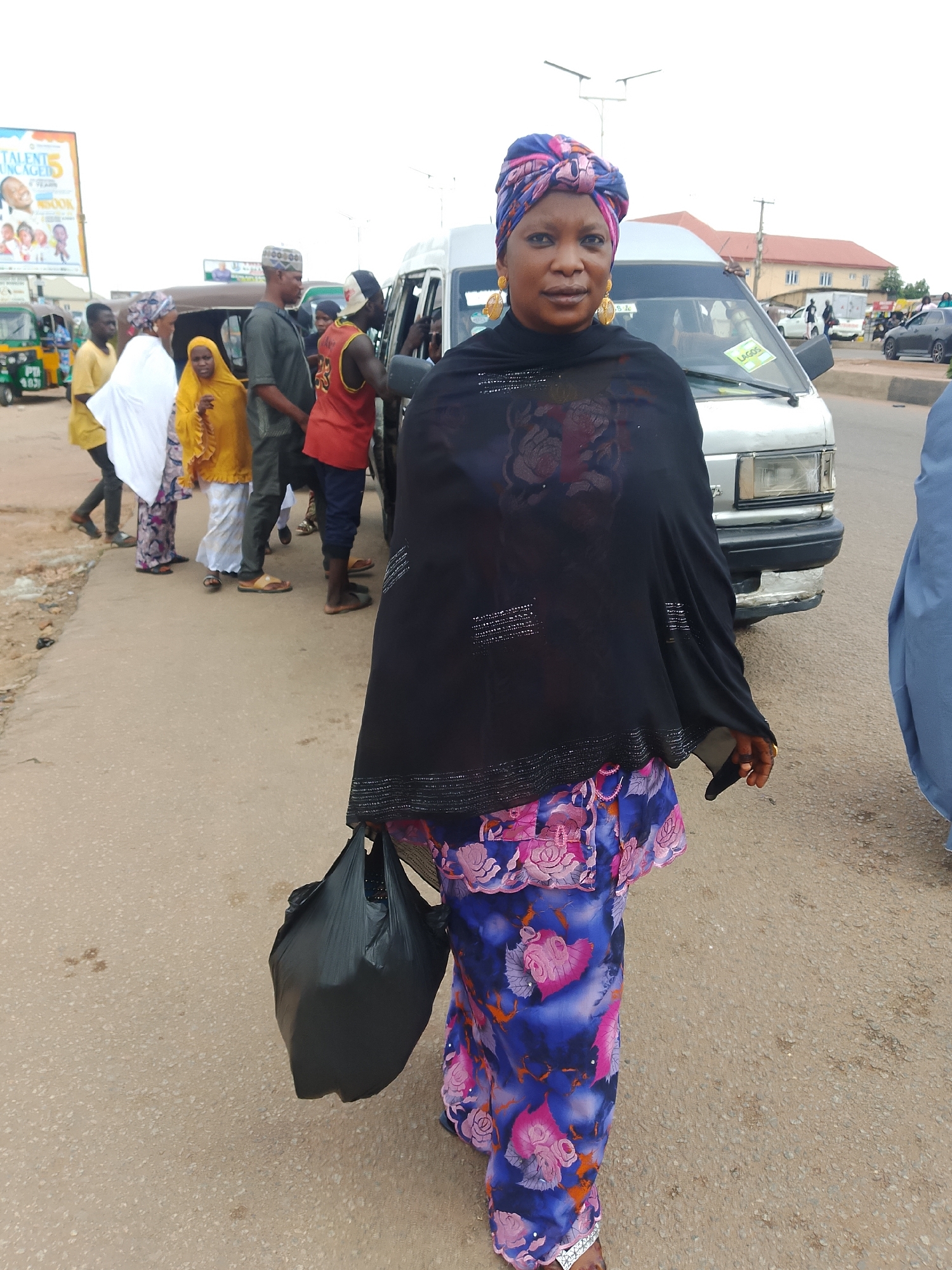 A woman in a black headscarf and a colorful blue and pink patterned skirt walks along a dusty road, carrying a black plastic bag in her right hand. She wears large gold hoop earrings. Behind her, other people are gathered near a white minivan and a green and yellow tricycle. A billboard with text that reads "TALENT UNLEASHED 5" is visible in the background to the left, along with buildings and utility poles. The scene appears to be a street in an urban area.