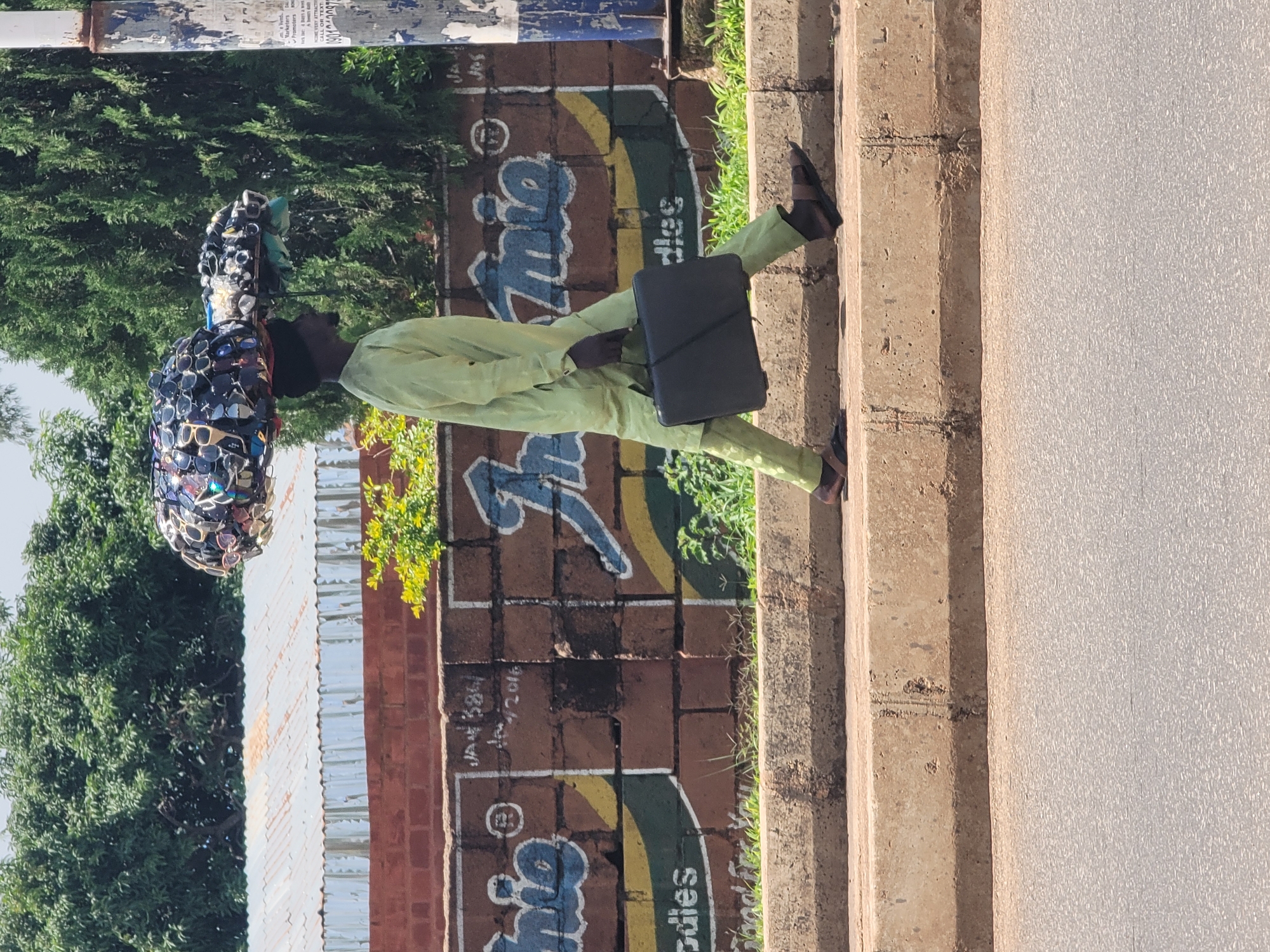 A man walks along a sidewalk in Barakin Lerawi, Nigeria. He is wearing a light green traditional outfit and carrying a black briefcase. On his head, he balances a large, round object covered in sunglasses. The man is walking away from the camera, towards a brick wall adorned with colorful graffiti. The wall is decorated with large blue letters and yellow and green curved shapes. A few green plants are growing near the wall. To the right of the man, concrete steps lead up to a textured, light-colored surface, possibly a wall or another building. The scene appears to be set during the daytime, with bright sunlight casting shadows. The overall atmosphere is ordinary, with the man engaged in his daily activity. There are no clear indicators of specific weather conditions beyond the presence of sunlight. The location cues are primarily the graffiti style and the presence of traditional attire, which are consistent with Nigerian settings.