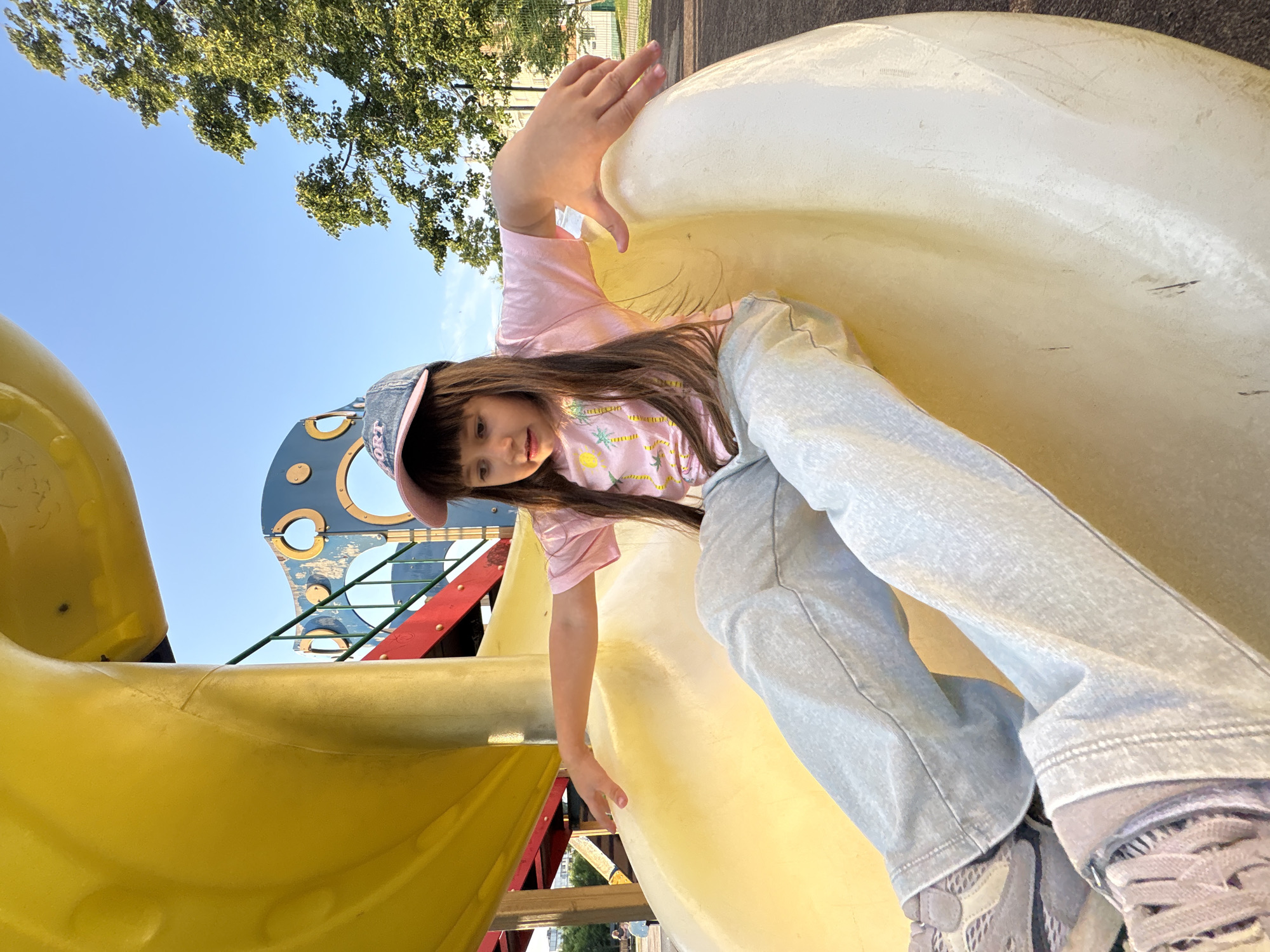 A young girl with long brown hair and a pink t-shirt, light blue jeans, and a blue and pink baseball cap sits on a yellow plastic slide at a playground. The girl is looking down and to her right, with her arms extended outwards. The slide appears to be part of a larger playground structure with yellow and red elements visible in the background. The sky is blue with some trees and buildings in the distance. This playground is located in the city of Chaikovsky, Russia.