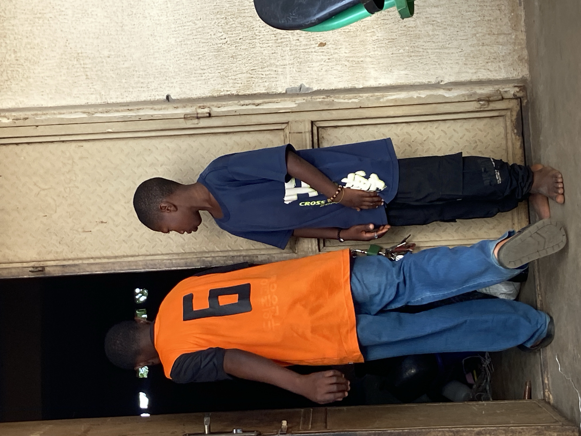 The image depicts two young males at a functional doorway in Mararaba, Nigeria. The figure on the left, an older boy, is seen from behind, in the process of entering or exiting a dark interior space. He wears an orange short-sleeved jersey with a black number '6' printed on the back, a black short-sleeved shirt underneath, blue denim jeans, and dark slip-on sandals. A keyring with multiple keys is attached near his right hip.
The figure on the right, a younger boy, stands barefoot in profile, facing left and observing the first boy. He wears a dark blue short-sleeved t-shirt with a vertical white graphic print that includes the word "CROSS" and other indistinct green/yellow elements. He has a multi-colored beaded bracelet on his left wrist and a ring on a finger of his left hand. His pants are dark-colored cargo-style trousers.
The doorway features two faded, light-tan metal doors with a diamond plate texture, both slightly ajar. The door on the left has a silver handle. The wall immediately to the right of the doors is a textured, light-colored plaster or concrete. The floor is light gray concrete, with a thin white cord visible horizontally in the foreground. A partial view of a green and black object, possibly a chair or stool, is present on the far right.