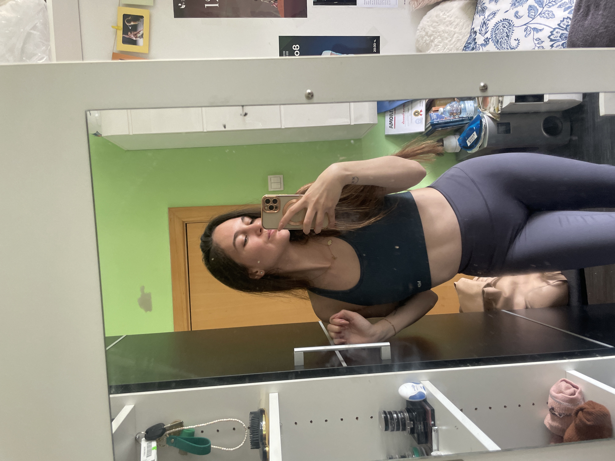 A woman is taking a selfie in a mirror. She is wearing a dark sports bra and gray leggings. The background includes a green wall, a brown door, a dresser with drawers open, and various items on the shelves of the dresser, including toiletries, keys, socks, and camera equipment. Based on the style of the room and the items present, the location is likely a residential interior. There is no indication in the image to confirm the location is Sofia, Bulgaria.