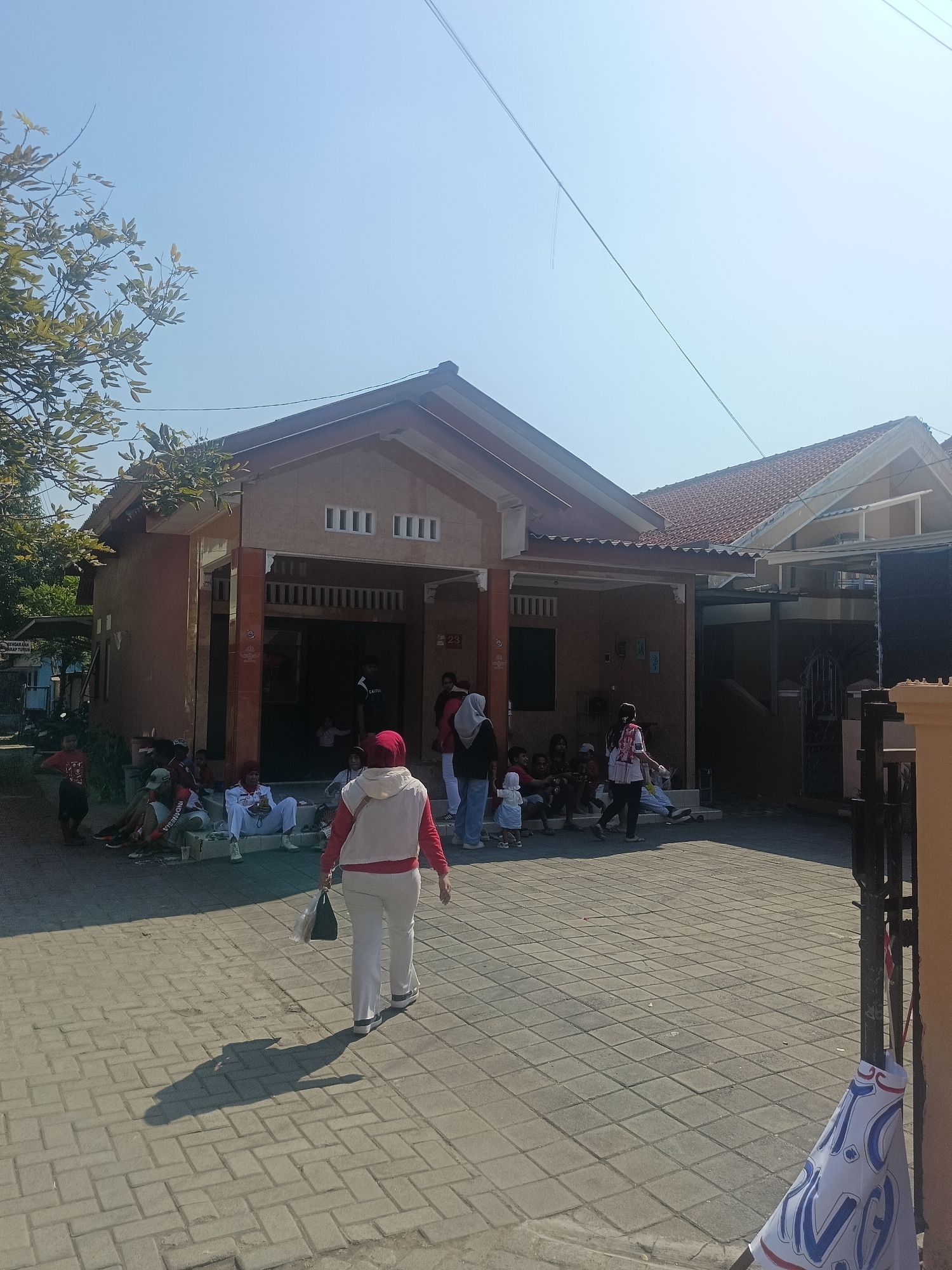 In Gedangan, Indonesia, on a clear, sunny day, a group of people are gathered outside a modest, single-story building with an orange facade and a tiled roof. The building appears to be a residence or a small community center. The ground in front of the building is paved with light gray rectangular interlocking bricks. Several individuals are seated or standing on the steps leading up to the building's entrance, while others are dispersed on the paved area. A woman wearing a red hijab and a white vest over a red shirt, along with white pants, walks away from the camera towards the right side of the frame, carrying a green bag. Behind her, to the left, a tree with green leaves casts a shadow on the pavement. A few children are visible among the seated groups, some appearing to be engaged in conversation or observation. A banner with blue and red lettering is partially visible on the right side of the frame, attached to a dark metal gate. The environment suggests a casual, possibly social or religious gathering in a residential area.