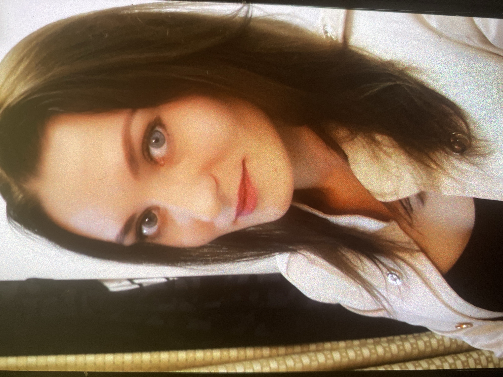 The media file presents a close-up, portrait-style photograph of a young woman with medium-long, dark brown hair. She has fair skin, light-colored eyes (appearing blue or gray), and a gentle, slight smile on her lips, which are colored with red or pink lipstick. Her gaze is directed straight at the viewer.
She is wearing a light-colored, possibly off-white or beige, collared shirt or jacket over a dark top. Visible details on her outerwear include a silver-toned button near her shoulder and a gold-toned button lower down.
The background is softly blurred, indicating an indoor setting. A dark, indistinct area is visible in the lower part of the frame, possibly furniture or another object. In the bottom right corner, a patterned fabric or surface is discernible, featuring a repeating motif of stylized letters that resemble a brand monogram, potentially "DIOR," although the text is somewhat blurry.
The woman's expression is calm and poised. The image captures a posed moment, without depicting any specific activity beyond being photographed. The lighting is soft and even. No explicit location cues are visible within the image itself, and the time of day or weather conditions cannot be determined. The contextual location provided is Saint Petersburg, Russia.