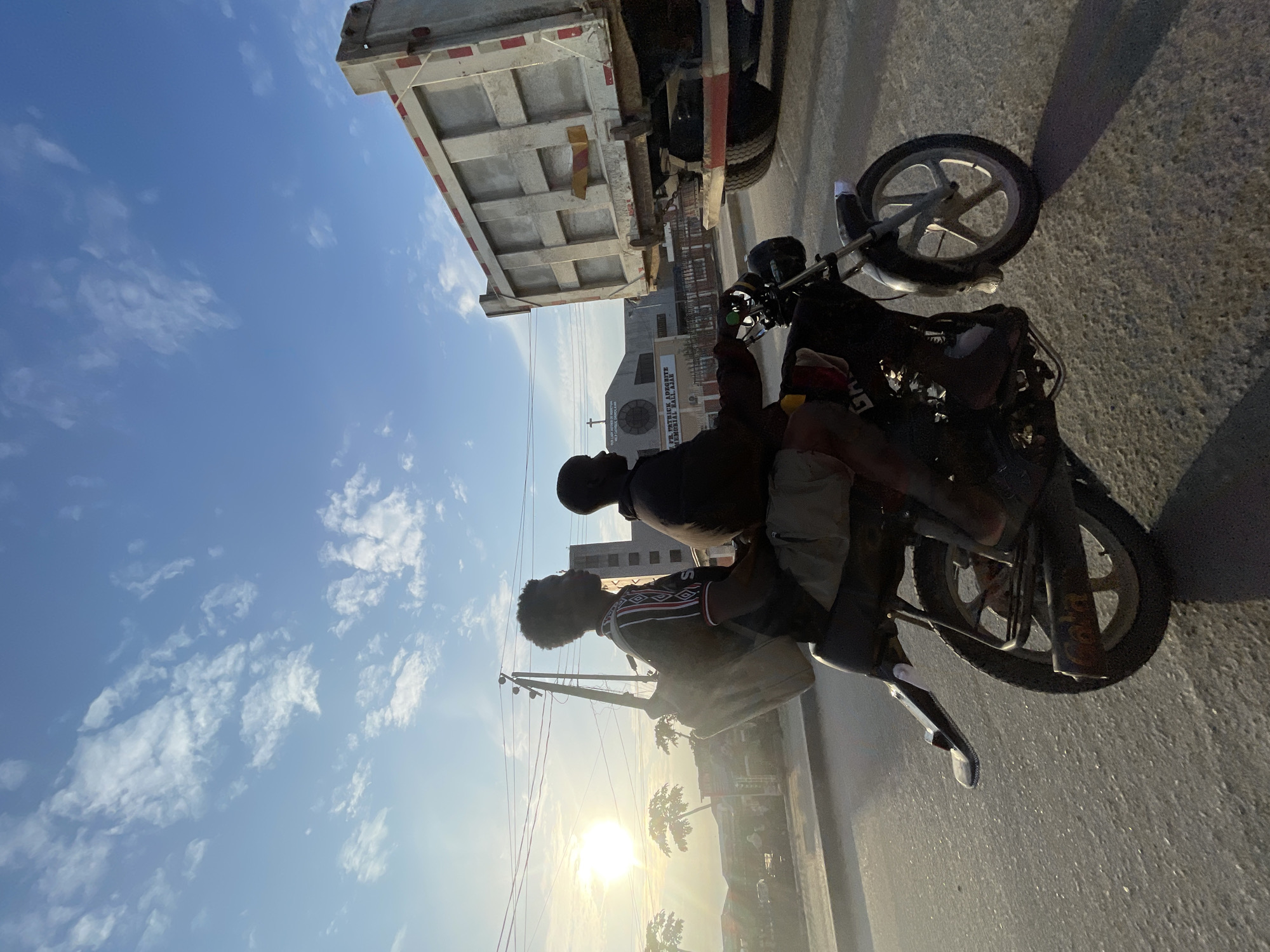 Two people are riding on a motorcycle. The person in front is driving, and the person behind is a passenger. They are both wearing casual clothing. The motorcycle is black and appears to be an older model.
The scene is a street in Ado, Nigeria. A large dump truck is parked on the side of the road. In the background, there are buildings and trees. The sun is setting, casting a warm glow over the scene. The sky is blue with a few white clouds. The overall mood is peaceful and serene.
There is visible text on a building in the background that appears to read "CHURCH OF THE FIRSTBORN INDUSTRIAL". There is also text on the motorcycle, but it is not clearly legible.