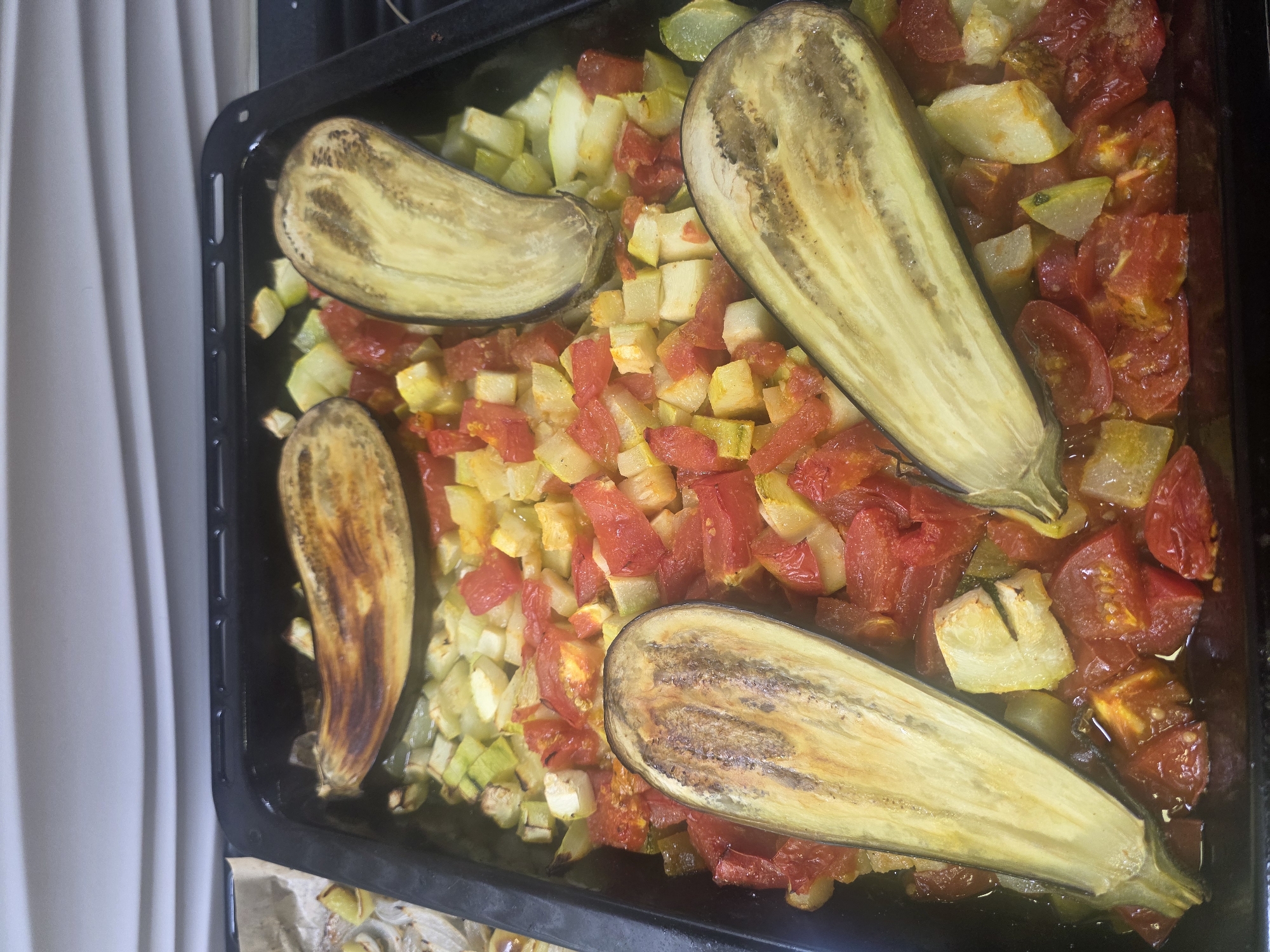 A close-up, overhead shot shows a black baking pan filled with roasted vegetables. The pan contains sliced eggplants, diced tomatoes, and chopped zucchini or squash. The eggplants are halved lengthwise and have visible grill marks and browning. The tomatoes appear soft and slightly caramelized, while the zucchini/squash pieces are lightly browned. The vegetables are glistening with oil, indicating they have been roasted. The background is out of focus and appears to be a textured white surface, possibly a wall or backsplash. The scene is indoors, in a kitchen environment. No people are visible in the image. The location is identified as Koloskovo, Russia.