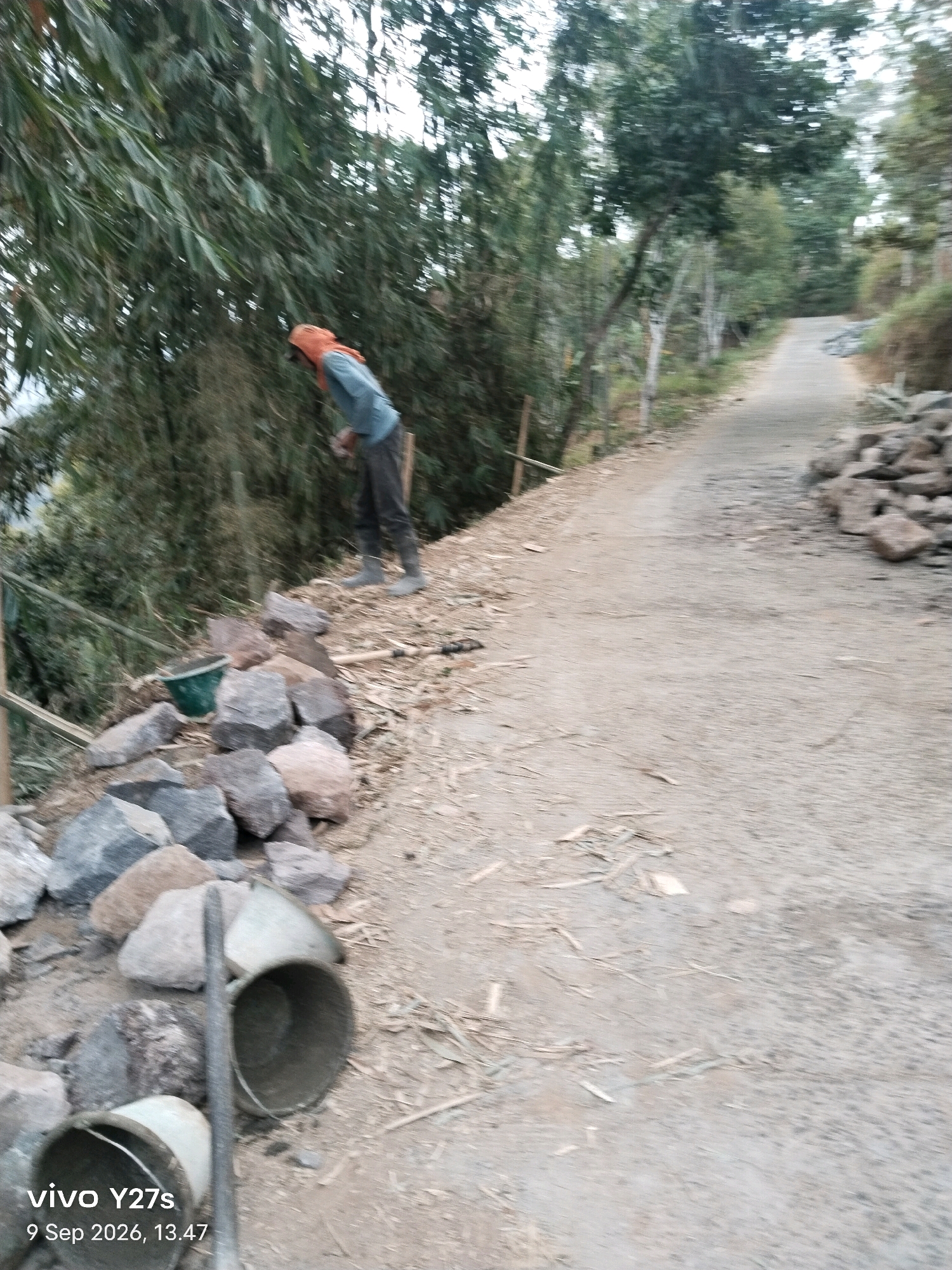 The image shows a rural scene with a person dressed in a blue shirt, dark pants, and gray boots, wearing an orange head covering. The person is standing on the side of a dirt road, facing away from the camera, and appears to be engaged in some sort of work involving construction materials. There are several large rocks and a green bucket near the person. The road curves to the right and ascends a gentle slope. Lush green vegetation, including bamboo, is visible on the left side of the road, with trees further up the slope. The overall setting suggests a construction or landscaping project in a hilly, natural environment. The bottom left corner of the image displays a watermark with "vivo Y27s" and "9 Sep 2026, 13.47". Based on the visual cues of the road and surrounding vegetation, this scene could plausibly be located in a rural area within Tamansari, Indonesia, known for its natural landscapes.