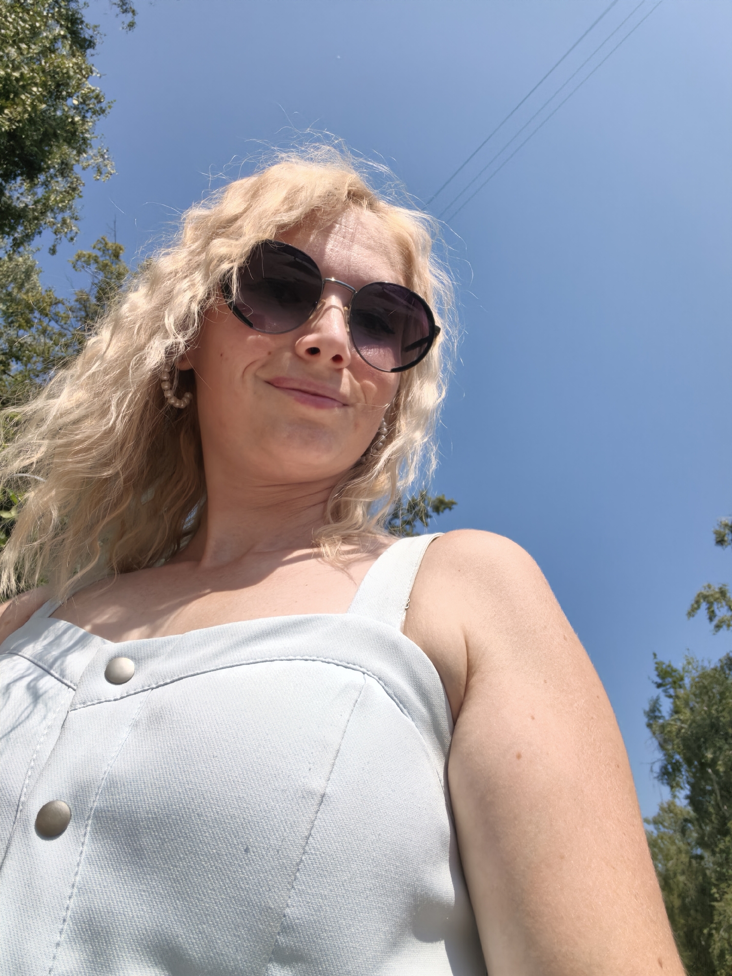 A low-angle, close-up, outdoor, daylight selfie shows a woman with blonde, curly hair and sunglasses looking down at the camera. She is wearing a light blue, sleeveless garment with two visible buttons. The background is a clear blue sky with some trees visible on the left and right sides. There are also power lines visible in the upper right portion of the image. The image was taken in Sterlitamak, Russia.