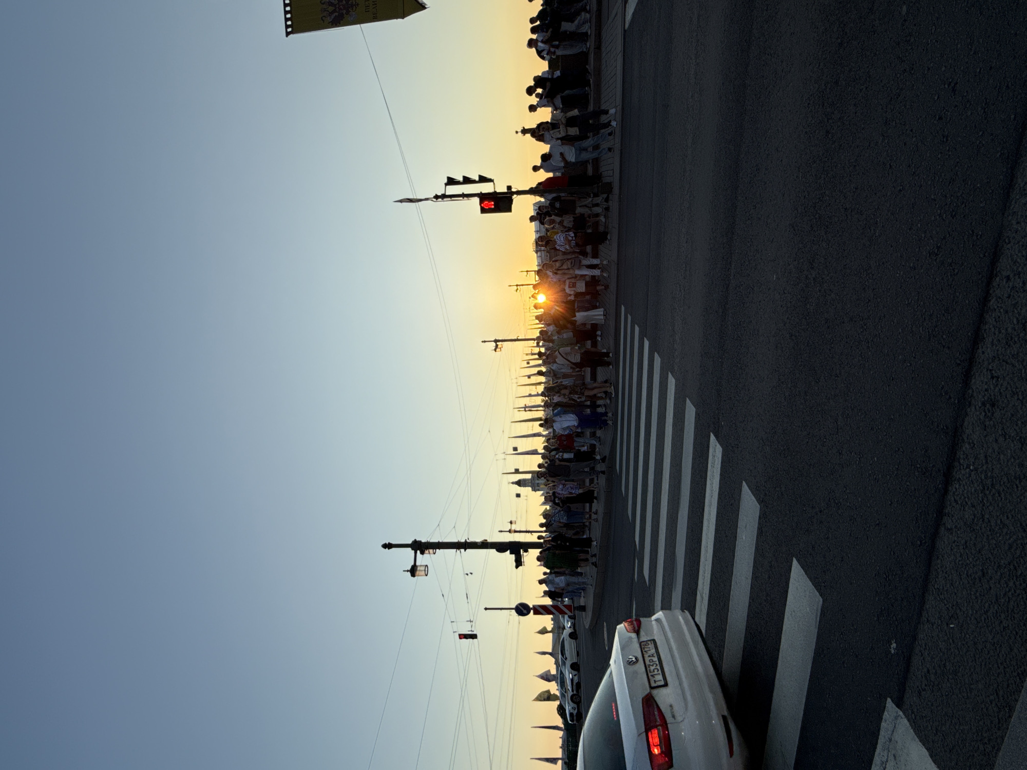 An outdoor, eye-level photograph captures a sunset scene on a clear evening in Saint Petersburg, Russia. In the foreground, a dark asphalt road with a white pedestrian crossing (zebra stripes) extends across the lower portion of the frame. A white Volkswagen Jetta sedan is partially visible on the far left, stopped with its rear bumper and license plate (T153PA 178, indicating Saint Petersburg region) positioned above the crossing.
In the midground, to the right of the pedestrian crossing, a large crowd of approximately 50-70 individuals is gathered on a sidewalk or embankment, mostly facing towards the brilliant setting sun on the horizon. Many people are silhouetted against the bright light, obscuring their details. To the left of the crowd, a tall black lamppost with multiple luminaires stands, equipped with a red traffic light facing the viewer. A circular "no entry" sign is visible behind it. Another red traffic light is visible further left.
The setting sun is positioned centrally on the horizon, casting a powerful golden-orange glare across the scene and reflecting intensely at eye level, making it difficult to discern distant features directly behind it. The sky transitions from a deep blue at the top to a warm orange and yellow near the horizon.
Along the embankment, behind the crowd, numerous flags are visible, appearing light-colored (possibly white), suggesting a festive or decorative arrangement. On the far right edge of the frame, a yellow banner with a dark double-headed eagle emblem and partially visible Russian text, "ПЕТР ВЕЛИКИЙ" (Peter the Great), hangs vertically. In the distant background, silhouetted against the bright sky, are faint outlines of buildings, potentially historical structures, suggesting the opposite bank of a wide river or bay. The specific location is identified as the **Palace Embankment (Dvortsovaya Naberezhnaya)**, looking west towards **Vasilyevsky Island** as the sun sets over the Neva River.