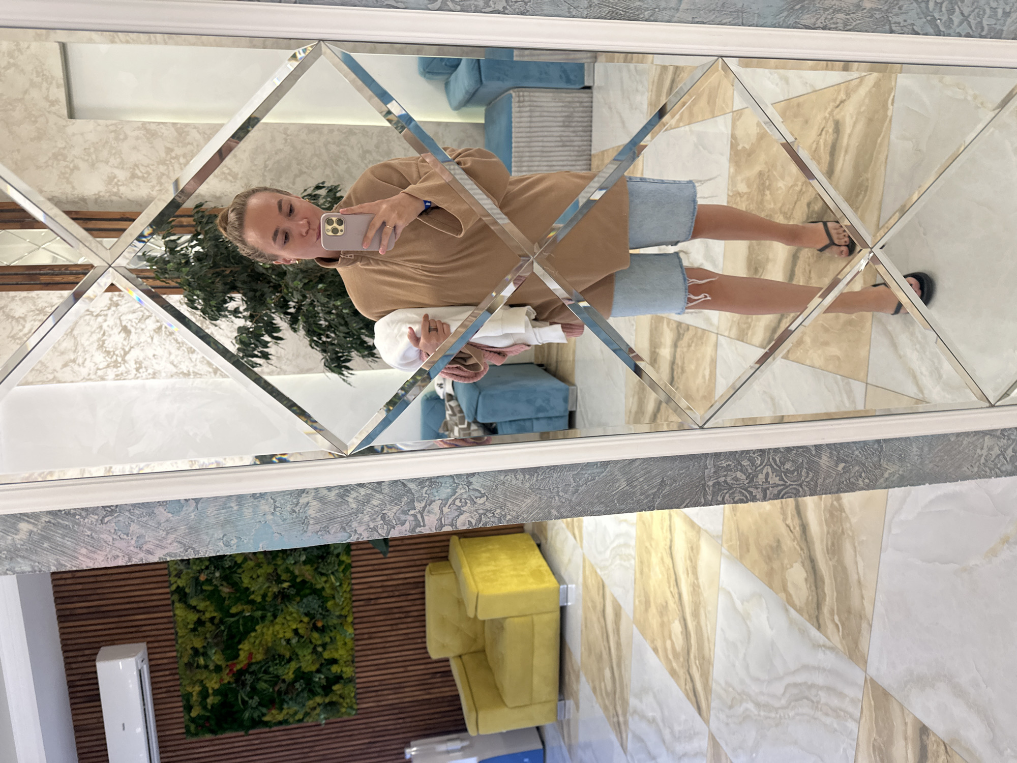 A woman is taking a mirror selfie in a room with modern decor. She is wearing a brown quarter-zip pullover and denim shorts. She is holding a white and pink garment in her left arm and her right hand is holding a smartphone. The room features a large, ornate mirror with a diamond pattern, a yellow armchair, blue seating, and a wall with vertical wood paneling and a green artificial plant installation. The floor is tiled with a marble-like pattern in shades of cream and beige. The scene suggests an indoor setting, possibly a hotel lobby or a boutique. The location of Vityazevo, Russia is not discernible from the image itself.