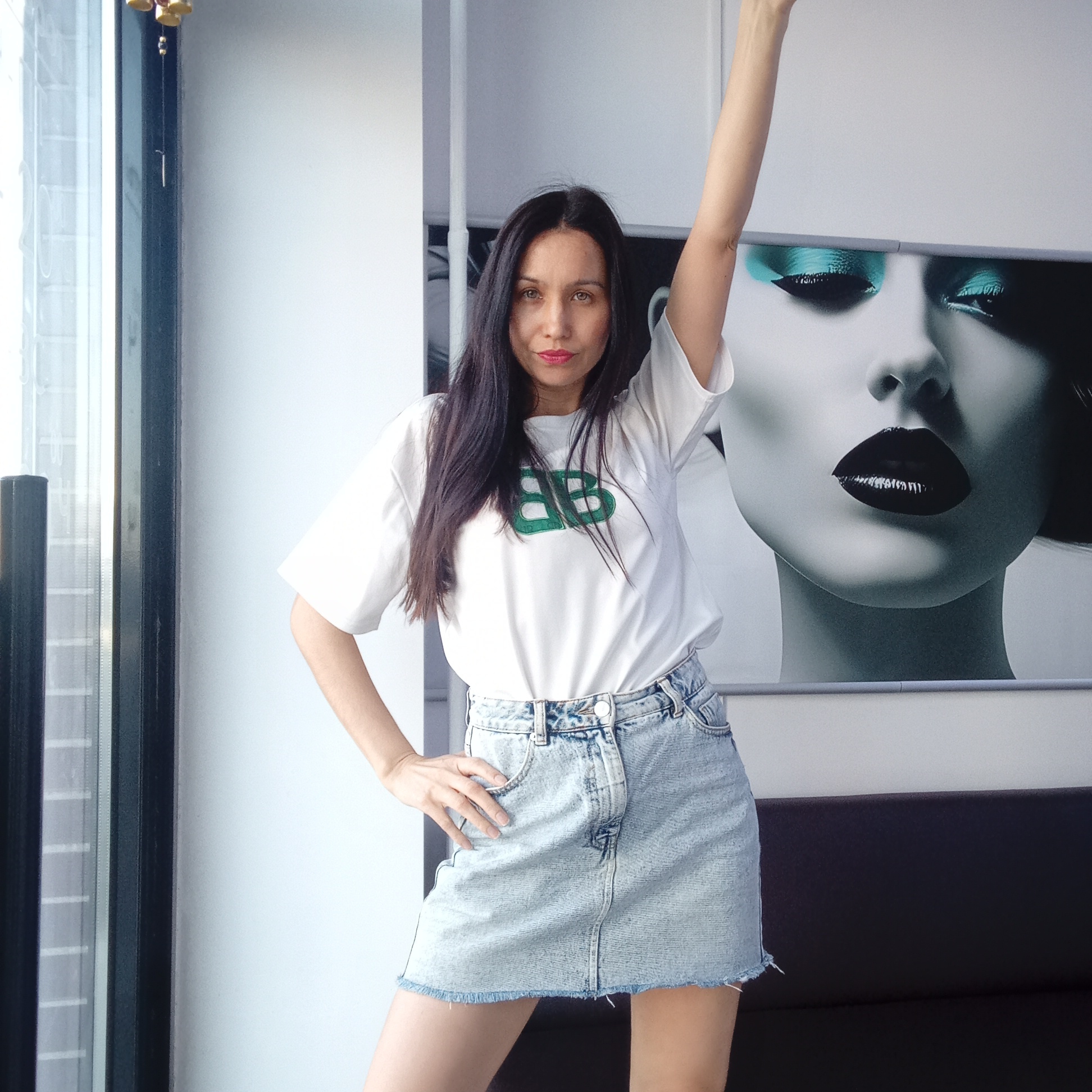 A woman with dark hair, wearing a white t-shirt with a green logo and a light blue denim skirt, is posing indoors. Her left hand is on her hip, and her right arm is raised. Behind her is a large black and white print of a woman's face with striking eye makeup. A window with a black frame is visible to the left, and a glimpse of gold-colored ornaments hanging in front of it can be seen. The scene appears to be in an interior setting within the city of Izhevsk, Russia.