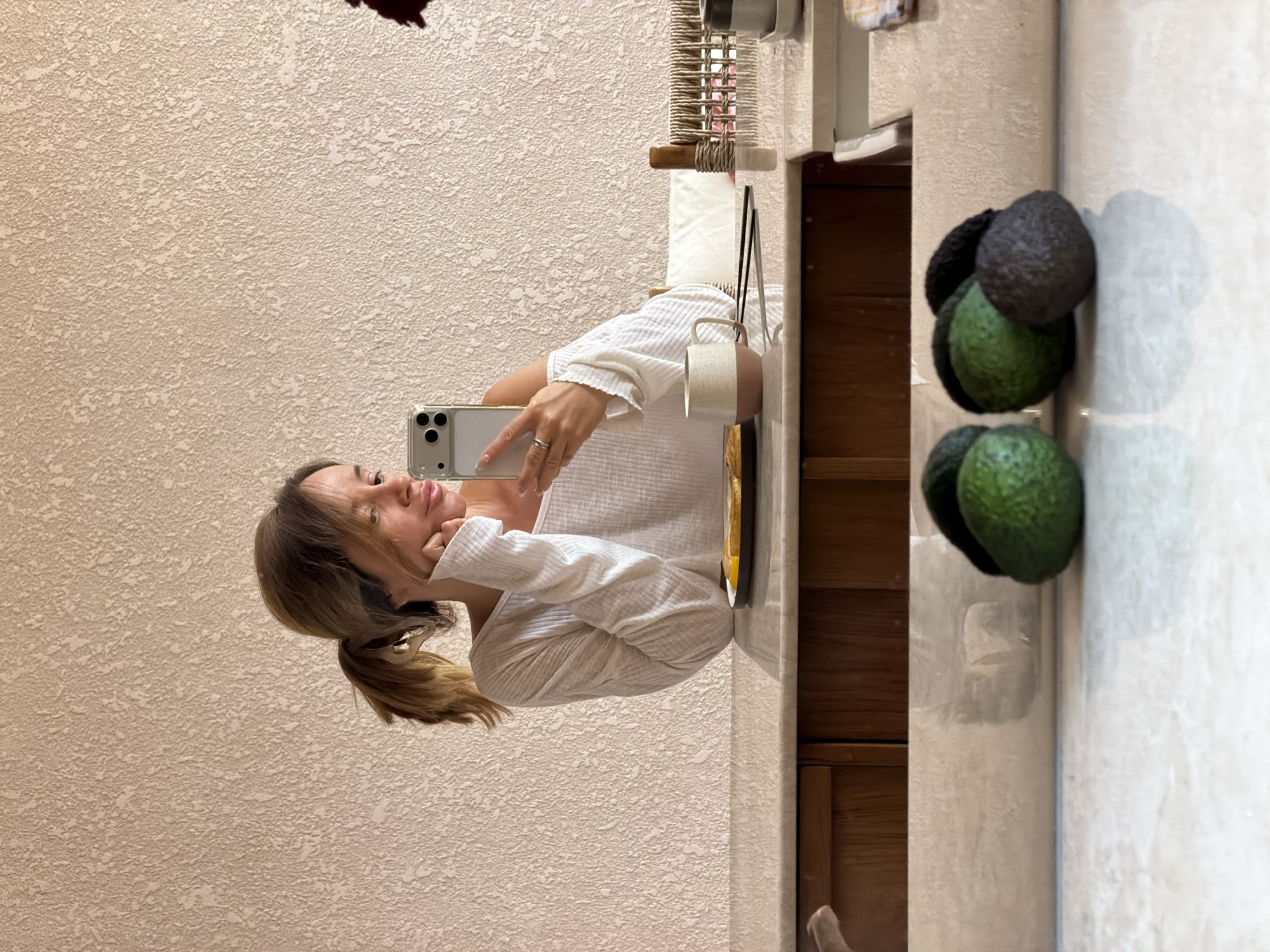 A woman is taking a selfie in a bathroom setting, which includes a cabinet, countertop, and decorative avocados. The scene is likely indoors and the specific location in Mengwi, Indonesia cannot be determined from the visual information.