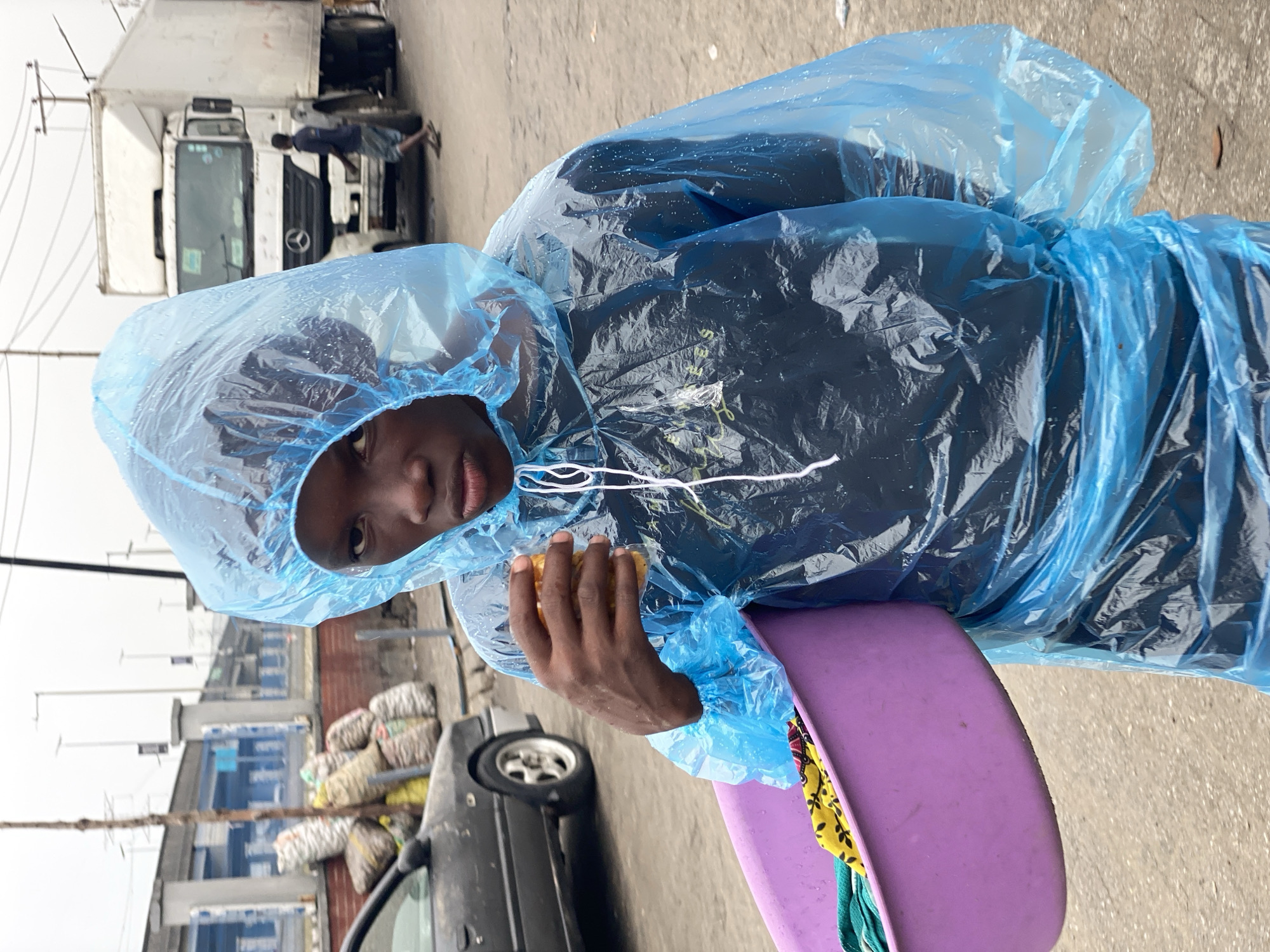 A young person in Lekki, Nigeria, wearing a light-blue plastic raincoat, looks directly at the camera. They hold a purple bowl and a piece of food in their hands. Behind the person are sacks of goods near a car, and further back, trucks are visible. The overall setting appears to be an outdoor marketplace.