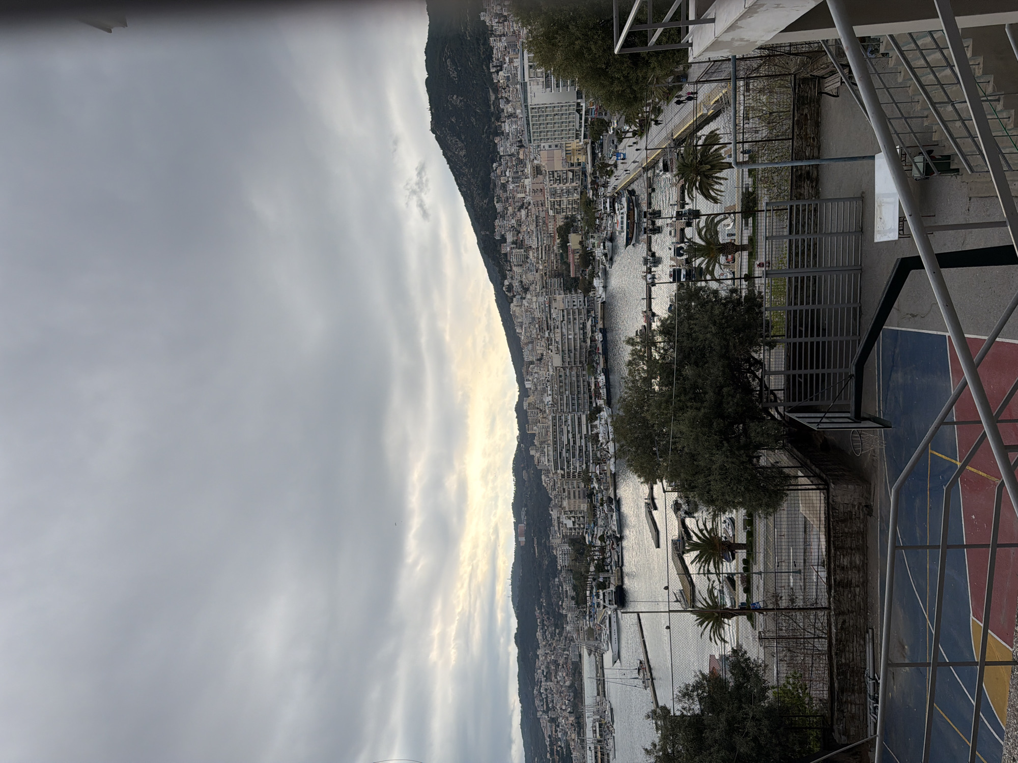 The image shows a view of Kavala, Greece, from an elevated position, possibly a balcony or rooftop. The city stretches along the coastline, with a backdrop of a mountain. The waterfront is visible, with numerous boats docked in the harbor. Buildings of various sizes and colors line the hillside. The sky is overcast with gray clouds. In the foreground, there is a sports court with a basketball hoop and fencing. Palm trees are visible along the waterfront.