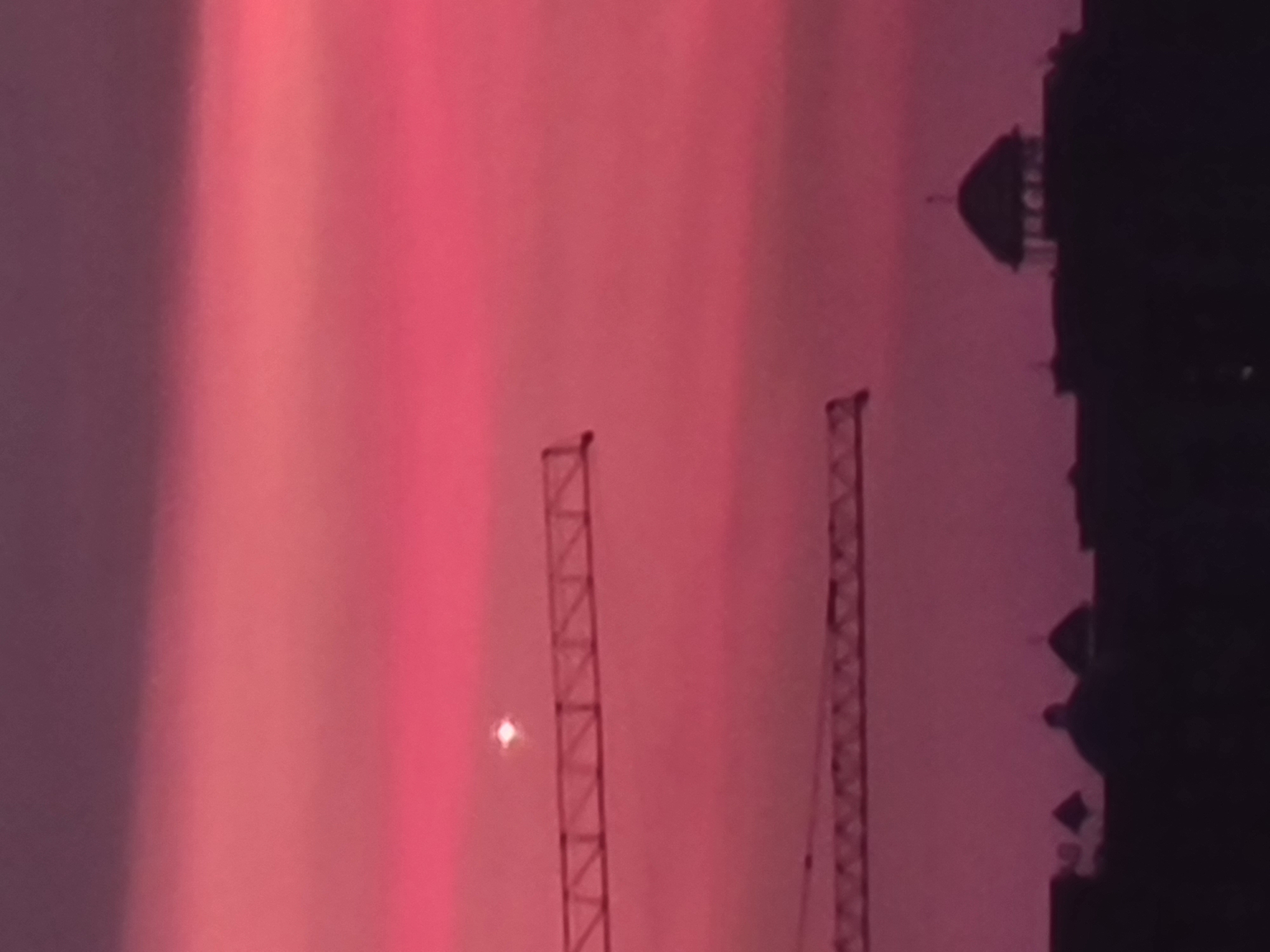 The image shows a construction site with two cranes against a pink sky at dusk. In the foreground, a silhouetted skyline of buildings and a domed structure is visible. A bright object, possibly a planet or a star, is present in the sky. The scene is likely located in Almaty, Kazakhstan, a city known for its construction and its proximity to the mountains, which can contribute to dramatic sky colors during sunrise and sunset. The overall impression is one of urban development juxtaposed with natural beauty.