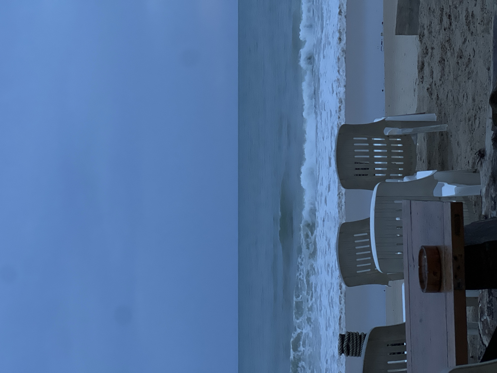 The image depicts a coastal scene on a possibly overcast day in the city of Unknown, Unknown. Dominating the right side of the frame are several white plastic chairs arranged near a wooden table. The table features a cylindrical object on top and appears to be set up on a sandy beach. The ocean, with visible waves, stretches out behind the beach area. The left side of the image is largely occupied by a cloudy, blue-toned sky.